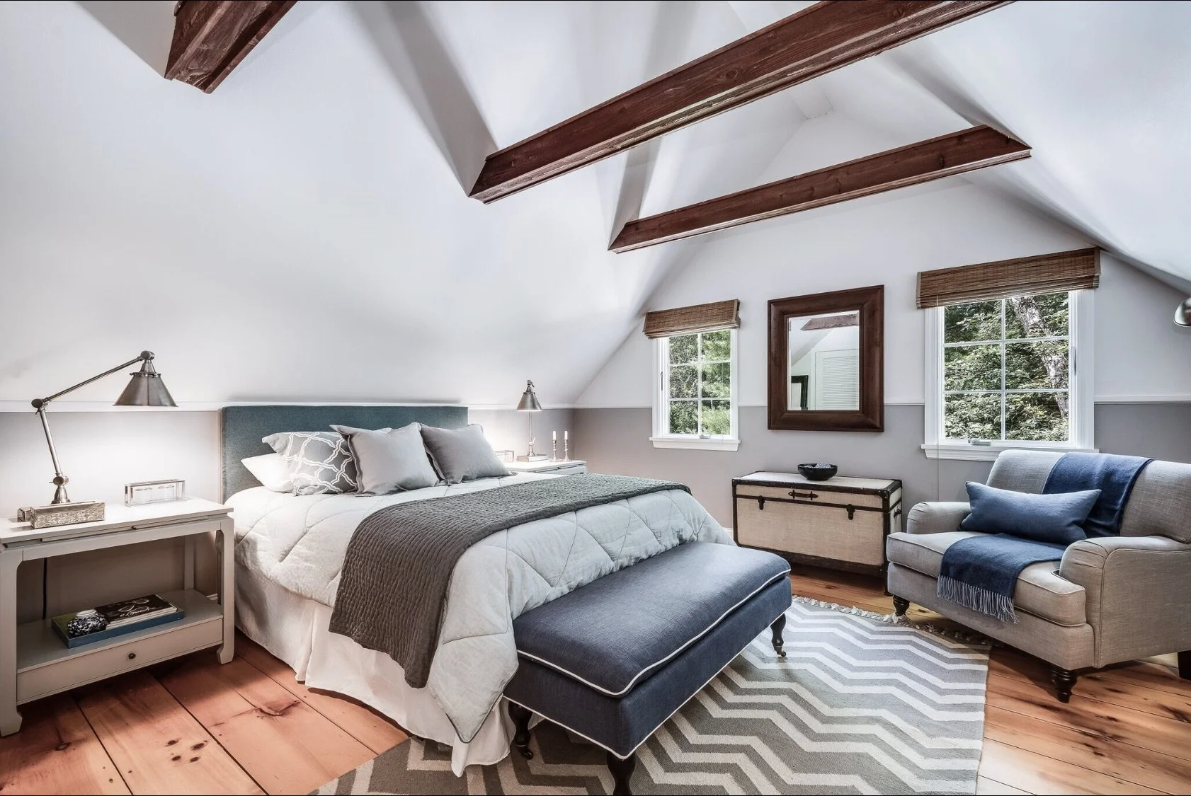 Cozy attic bedroom with exposed wood beams, soft neutral bedding, a patterned rug, and seating area beneath pitched ceilings
