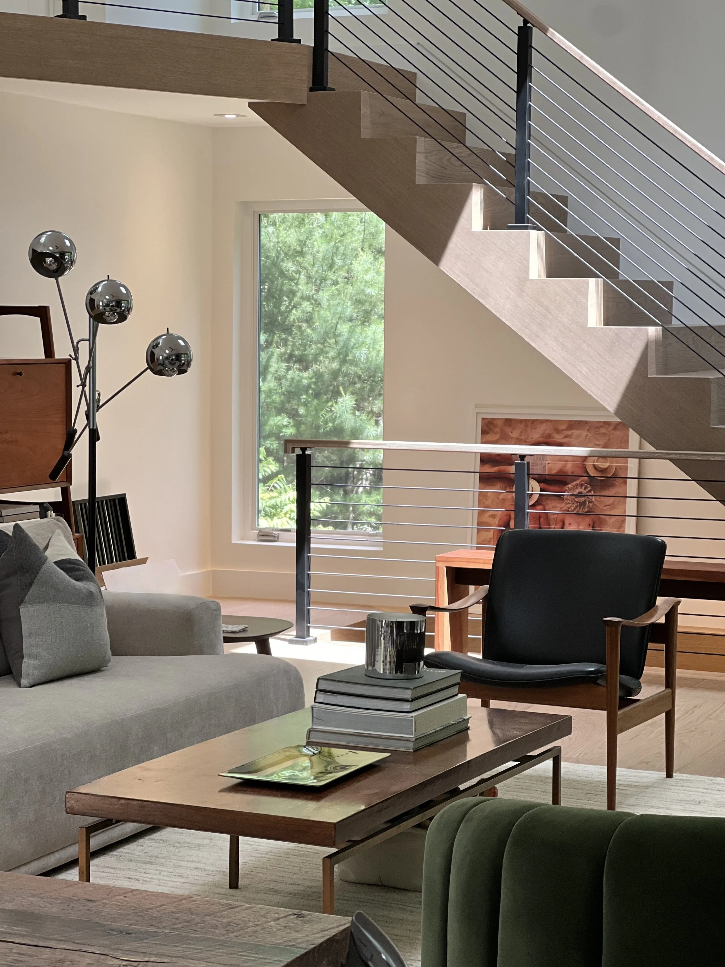 Open living space beneath a floating staircase, featuring neutral seating, wood accents, and metal railings with views to surrounding greenery