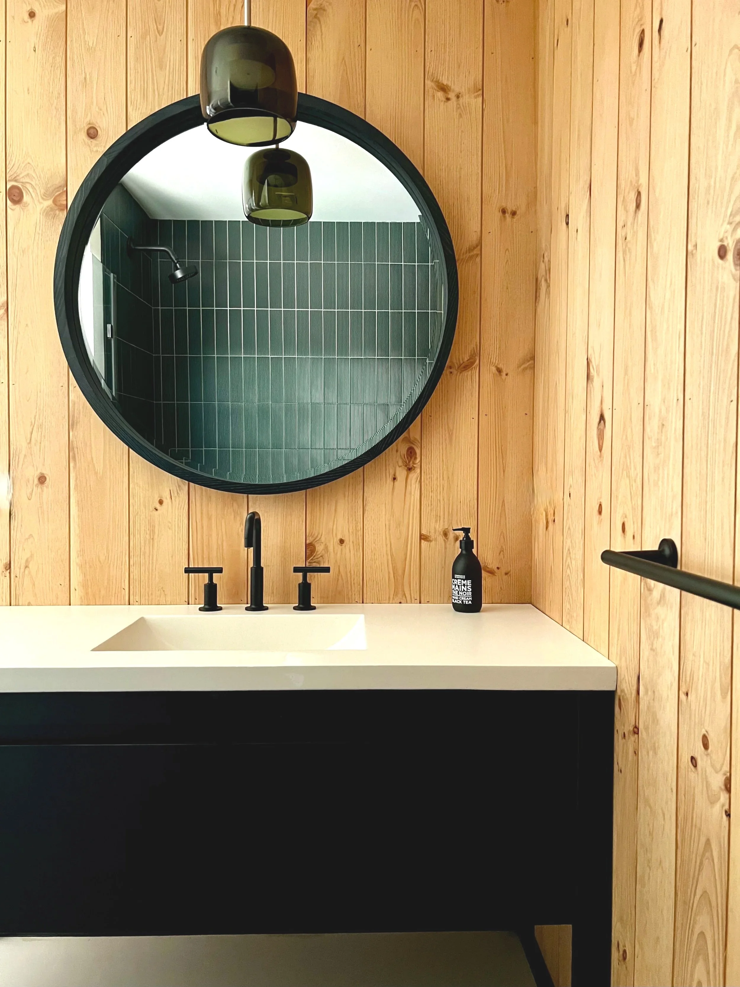 Clean modern bathroom with wooden wall paneling, large round black-framed mirror, white sink, black faucet and handles, a small black soap dispenser, and a black towel bar. Reflection shows green ceramic tiled shower with a black showerhead.