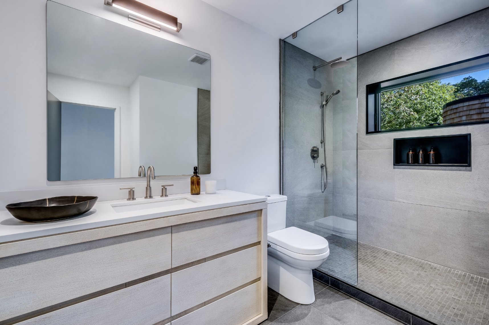 Contemporary bathroom with streamlined light wood vanity, integrated sink, glass shower enclosure, and neutral stone tile accents