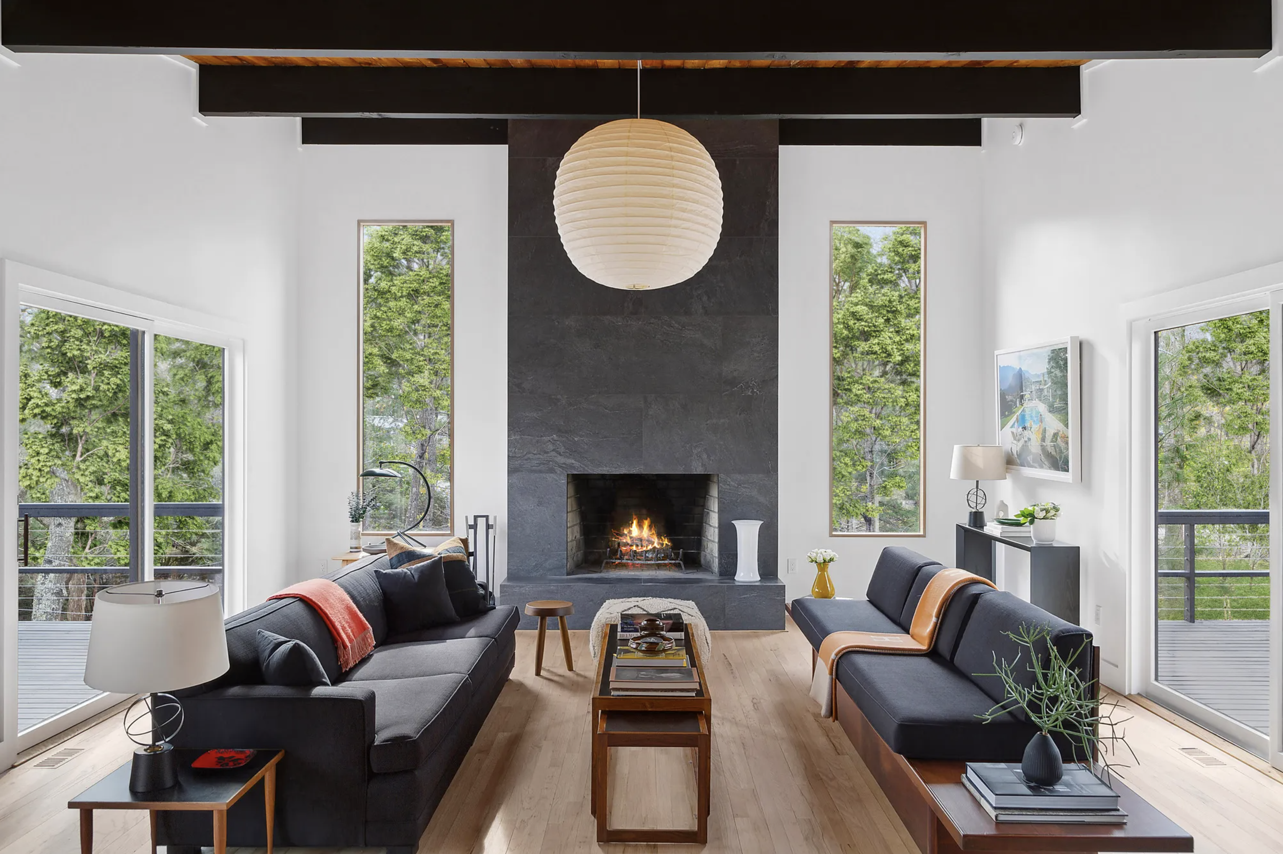 Living room with two dark sofas facing each other, a white fireplace with a burning fire at the center, large windows showing green trees outside, and a round hanging paper lantern ceiling light.