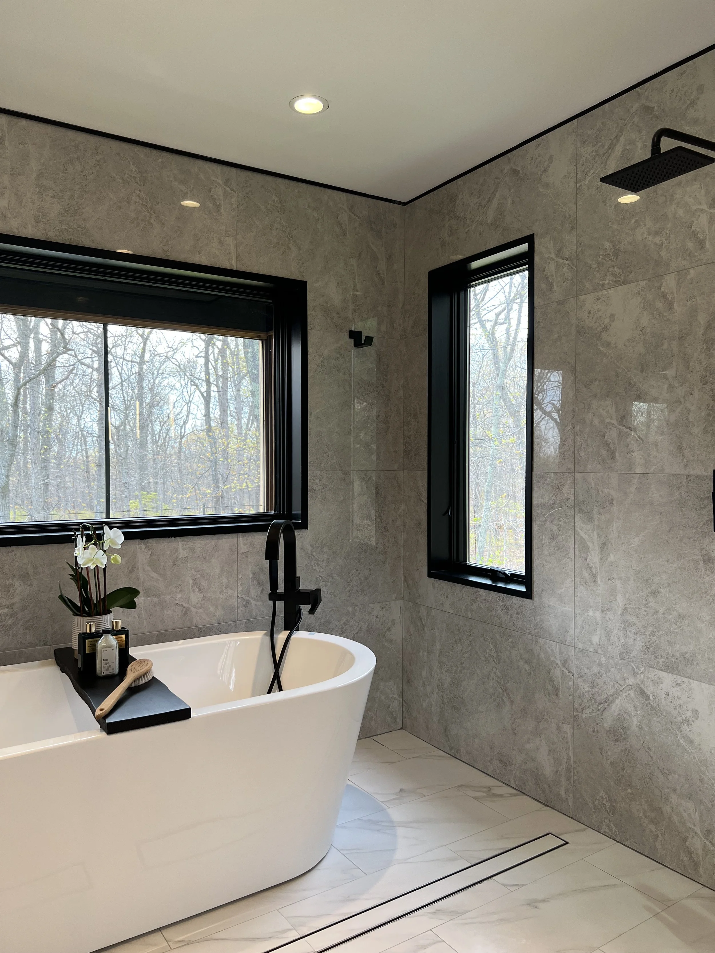 Spa-like bathroom with freestanding soaking tub, large gray stone tile walls, black fixtures, and woodland views through picture windows