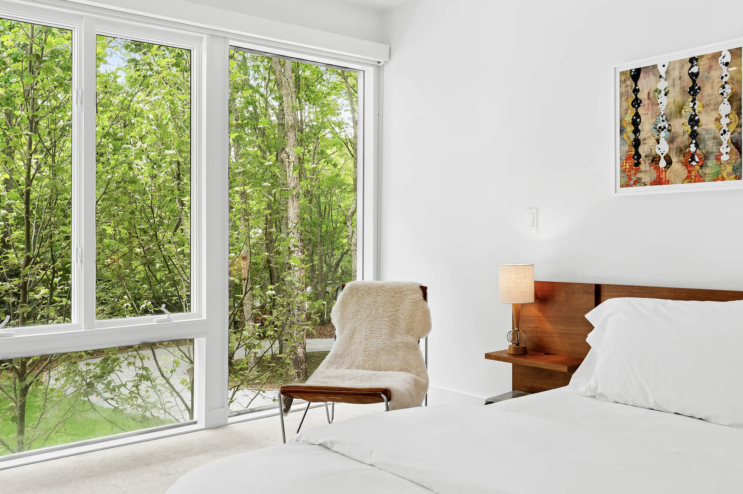 Bedroom sitting area with floor-to-ceiling windows, upholstered lounge chair, wood nightstand, and leafy outdoor views