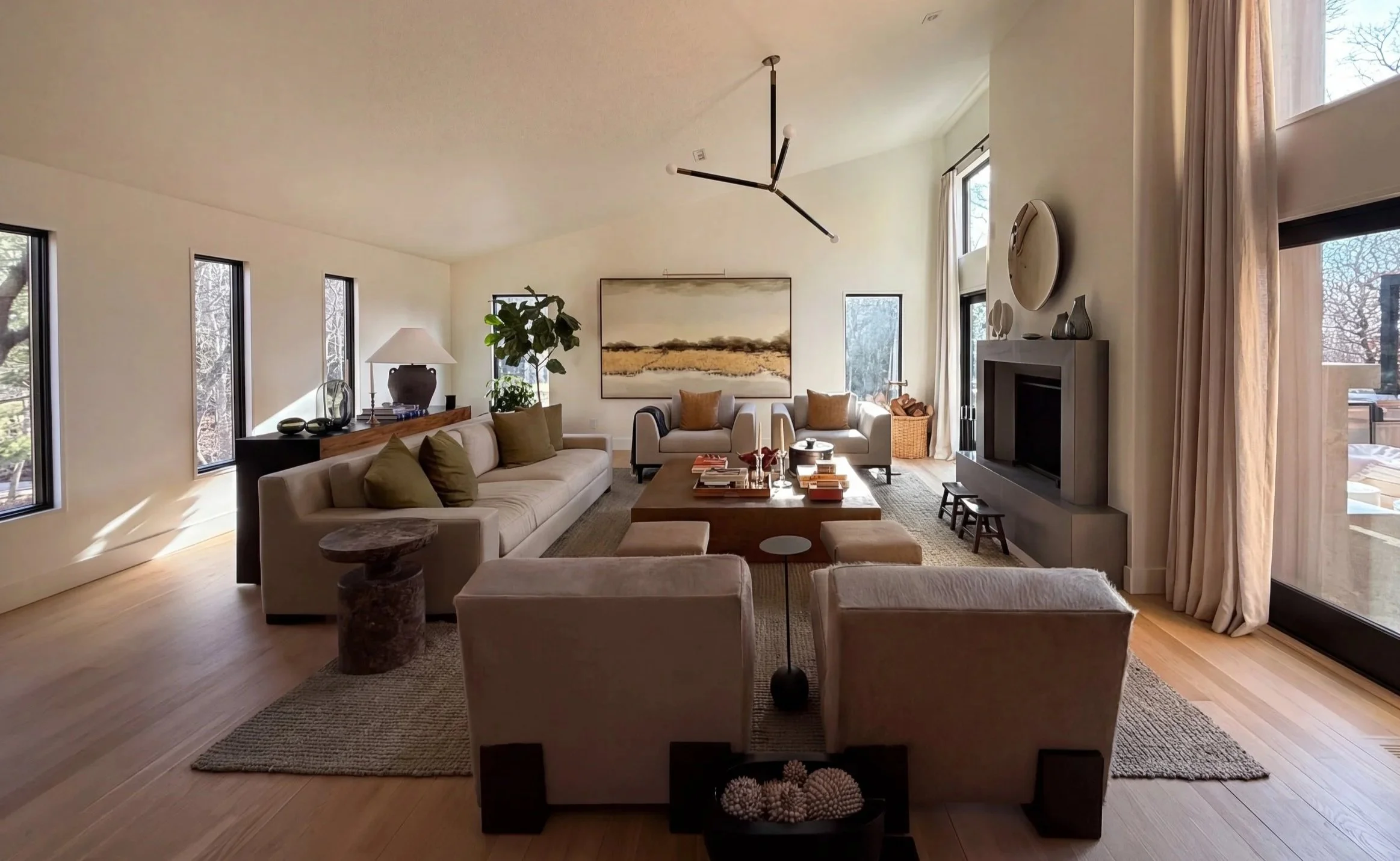 Light-filled living room with neutral seating arranged around a central coffee table, modern fireplace, sculptural lighting, and tall windows framing wooded views