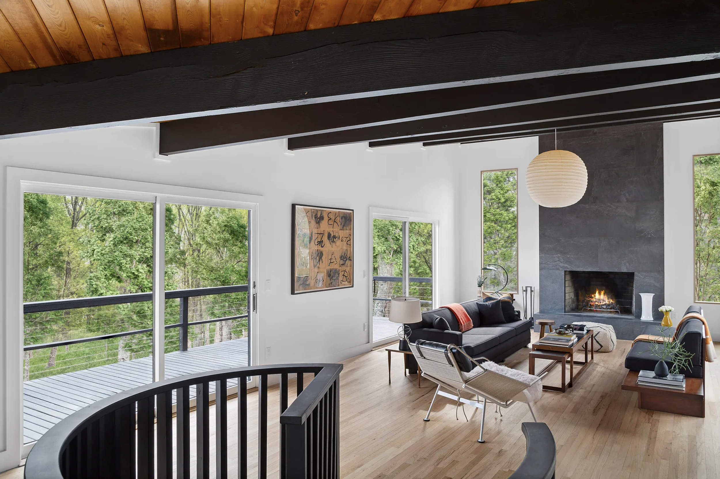 Open living area with exposed dark wood beams, light oak floors, modern seating, and large glass doors opening to a wooded deck