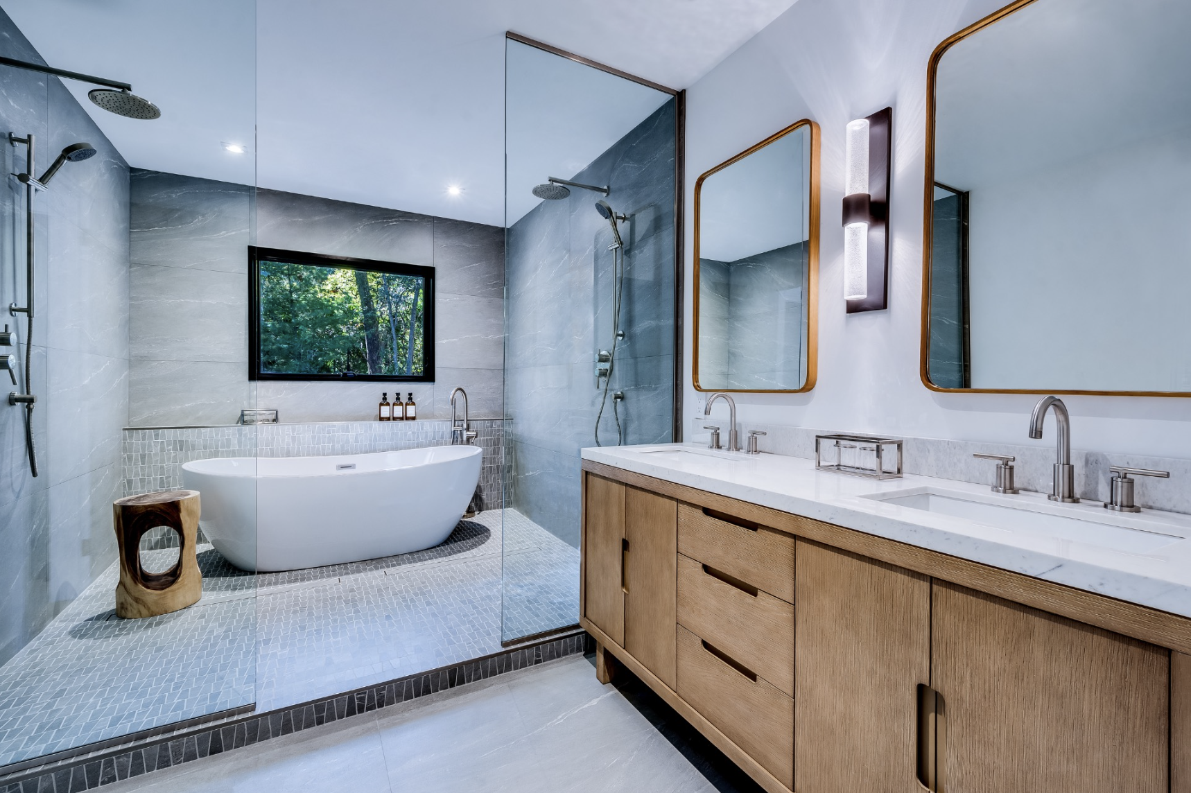 Spa-like bathroom with freestanding soaking tub, glass-enclosed shower, light wood double vanity, and soft gray stone tile throughout