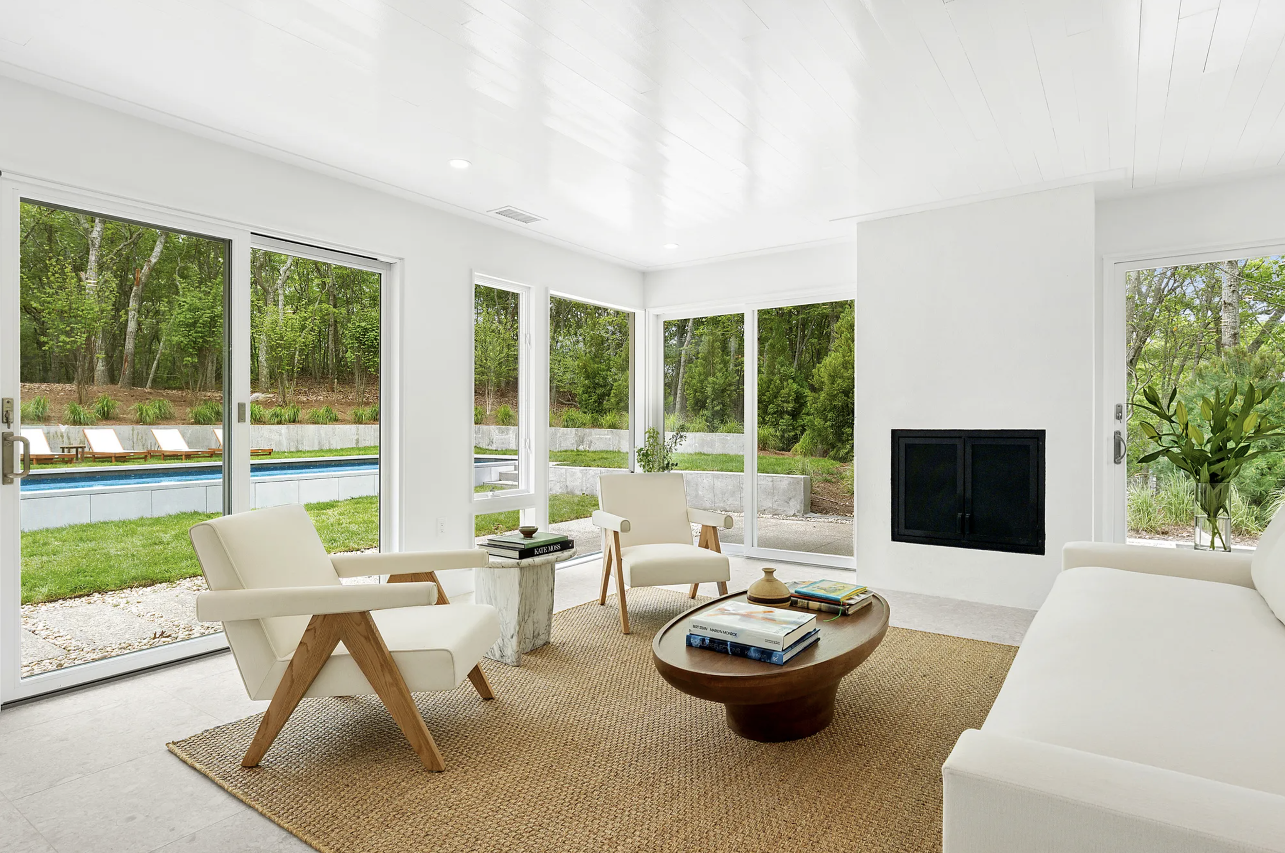 Light-filled living room with floor-to-ceiling glass doors, minimalist seating, indoor fireplace, and views to a landscaped backyard and pool