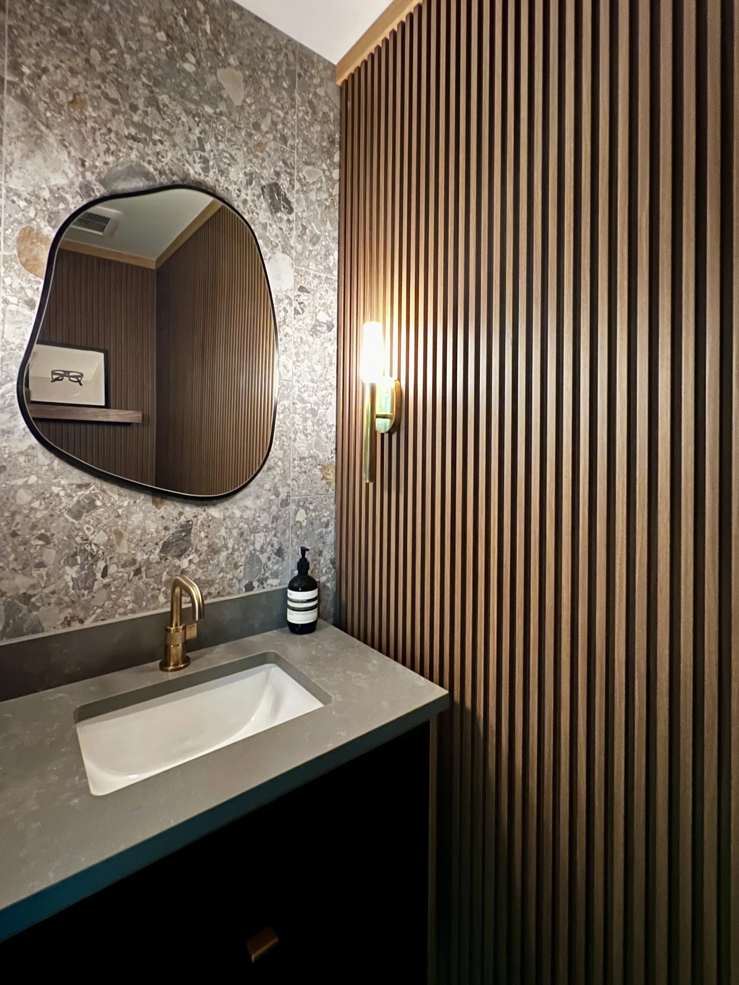 Moody powder room with stone backsplash, vertical wood slat wall, sculptural mirror, and a minimalist vanity with brass faucet