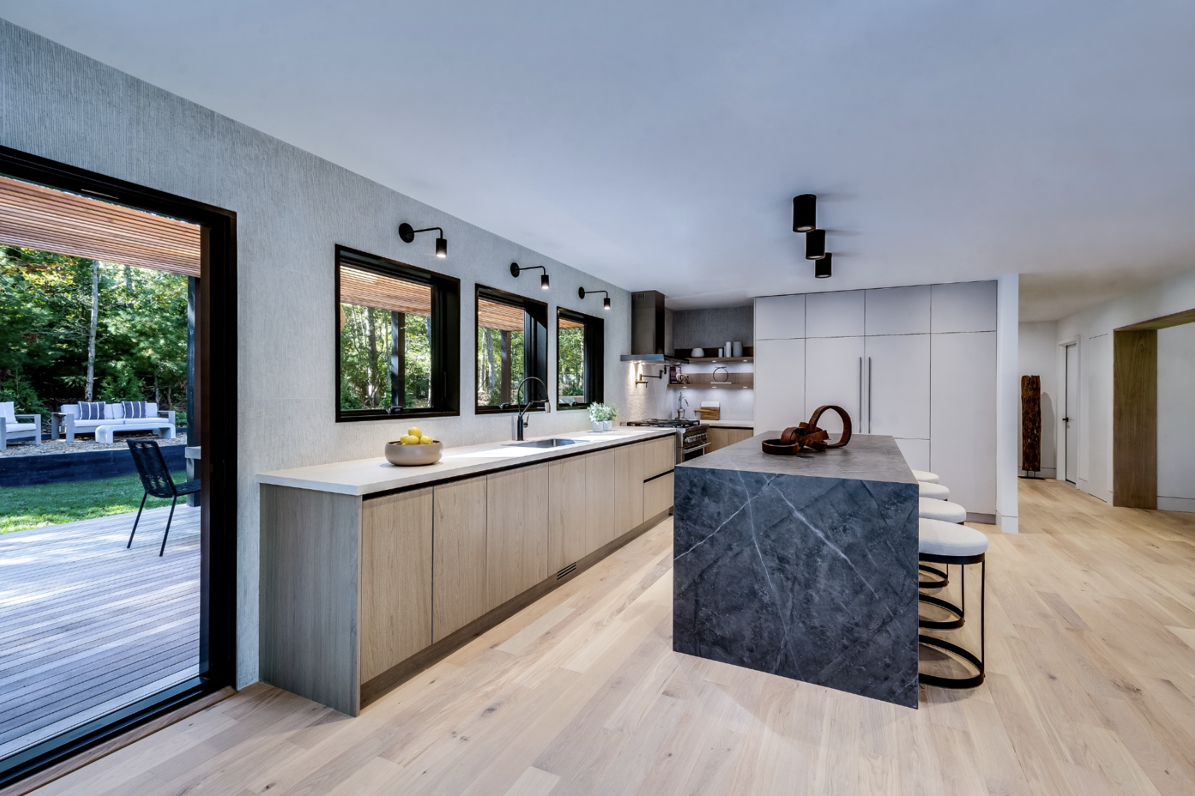 Open kitchen with light oak cabinetry, marble waterfall island, integrated seating, and large windows opening to a wooded outdoor deck