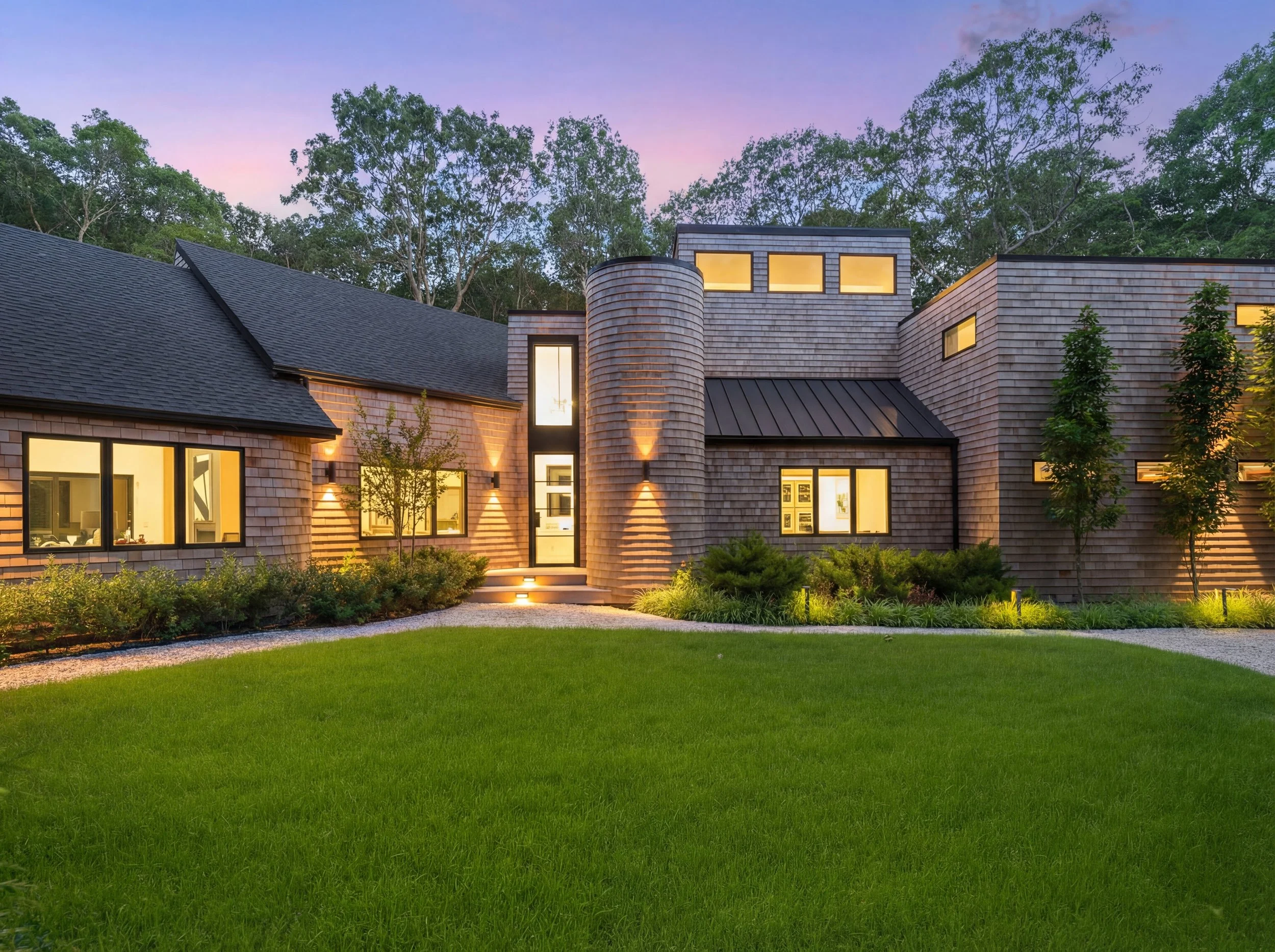 Exterior view of a cedar-shingled home with modern massing, illuminated windows, and landscaped lawn at dusk