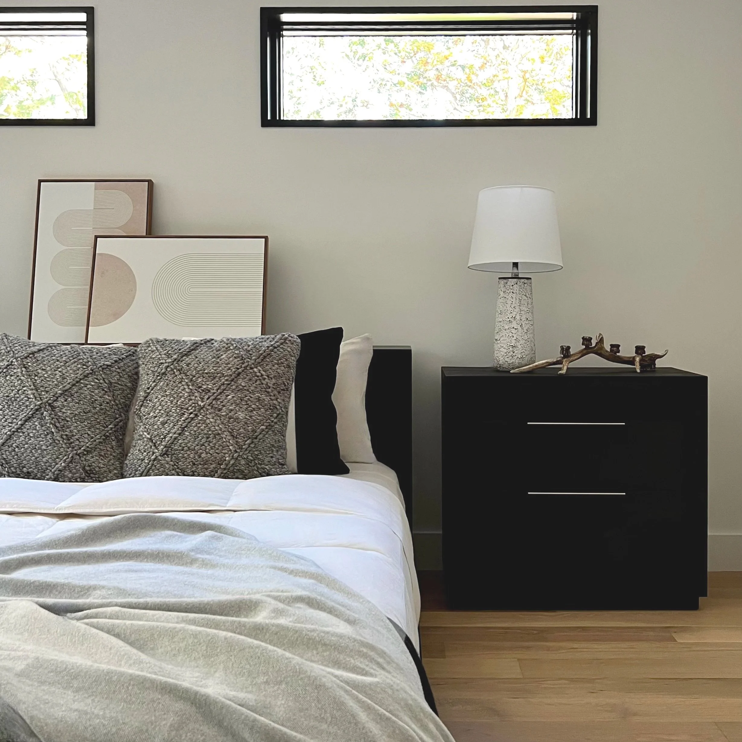 A bedroom with a bed having patterned pillows, black and white cushions, and geometric art prints on the wall. There is a black nightstand with a white table lamp, decorative branch, and two drawers. Two small rectangular windows are above the wall.