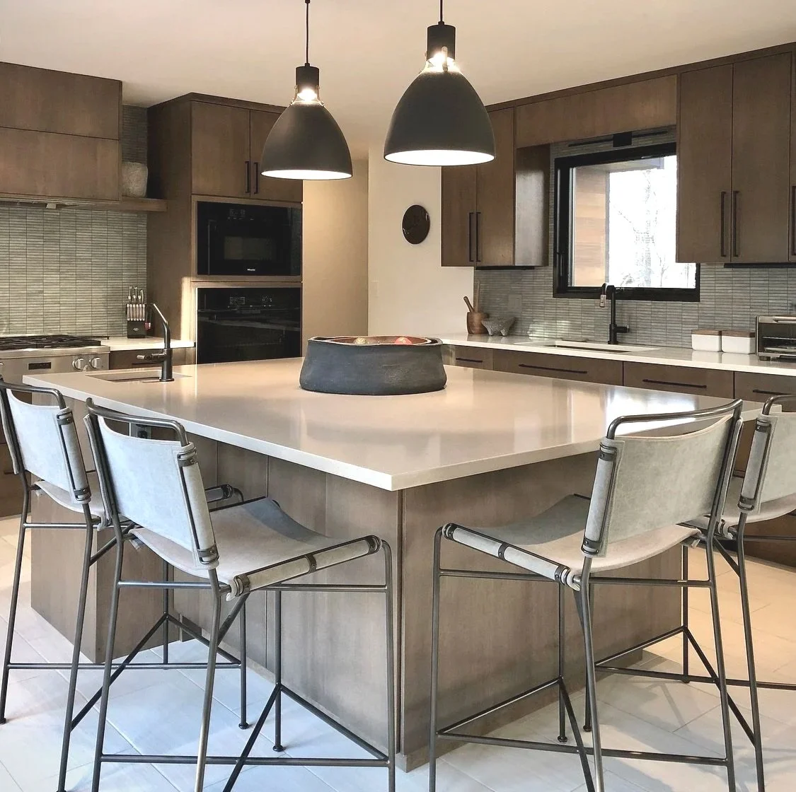 Modern kitchen with a large island, three hanging black pendant lights, and wooden cabinets. There are four metal chairs around the island, and a black bowl in the center. The kitchen has a window above the sink and built-in appliances.