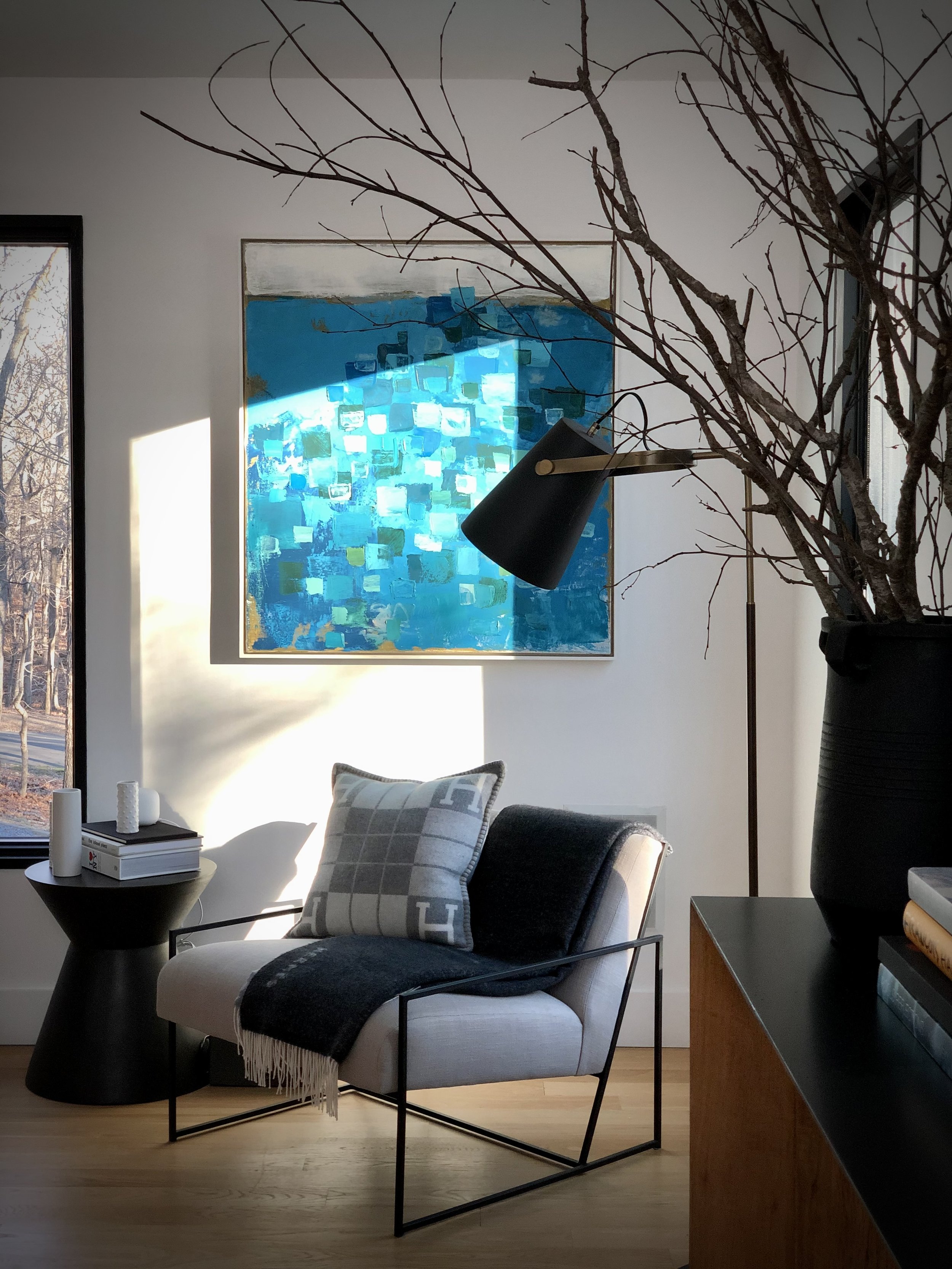 Reading corner with upholstered lounge chair, black metal floor lamp, abstract blue artwork by Michael Abramson, and sculptural branches in a tall vase