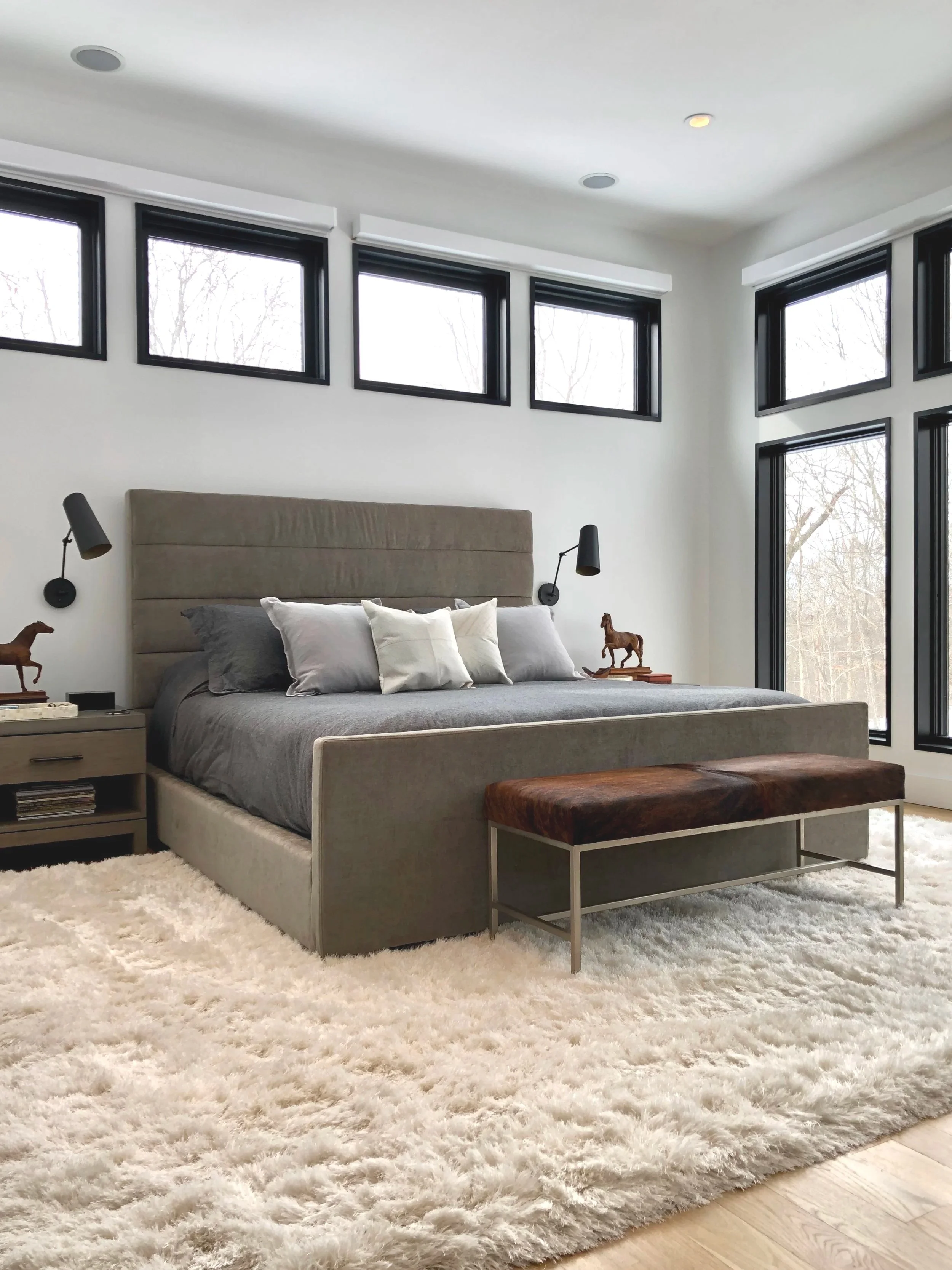 Modern bedroom with beige upholstered bed, gray bedding, white and gray pillows, black wall-mounted reading lamps, black-framed windows, a brown bench at foot of bed, beige nightstand, and a plush cream area rug.