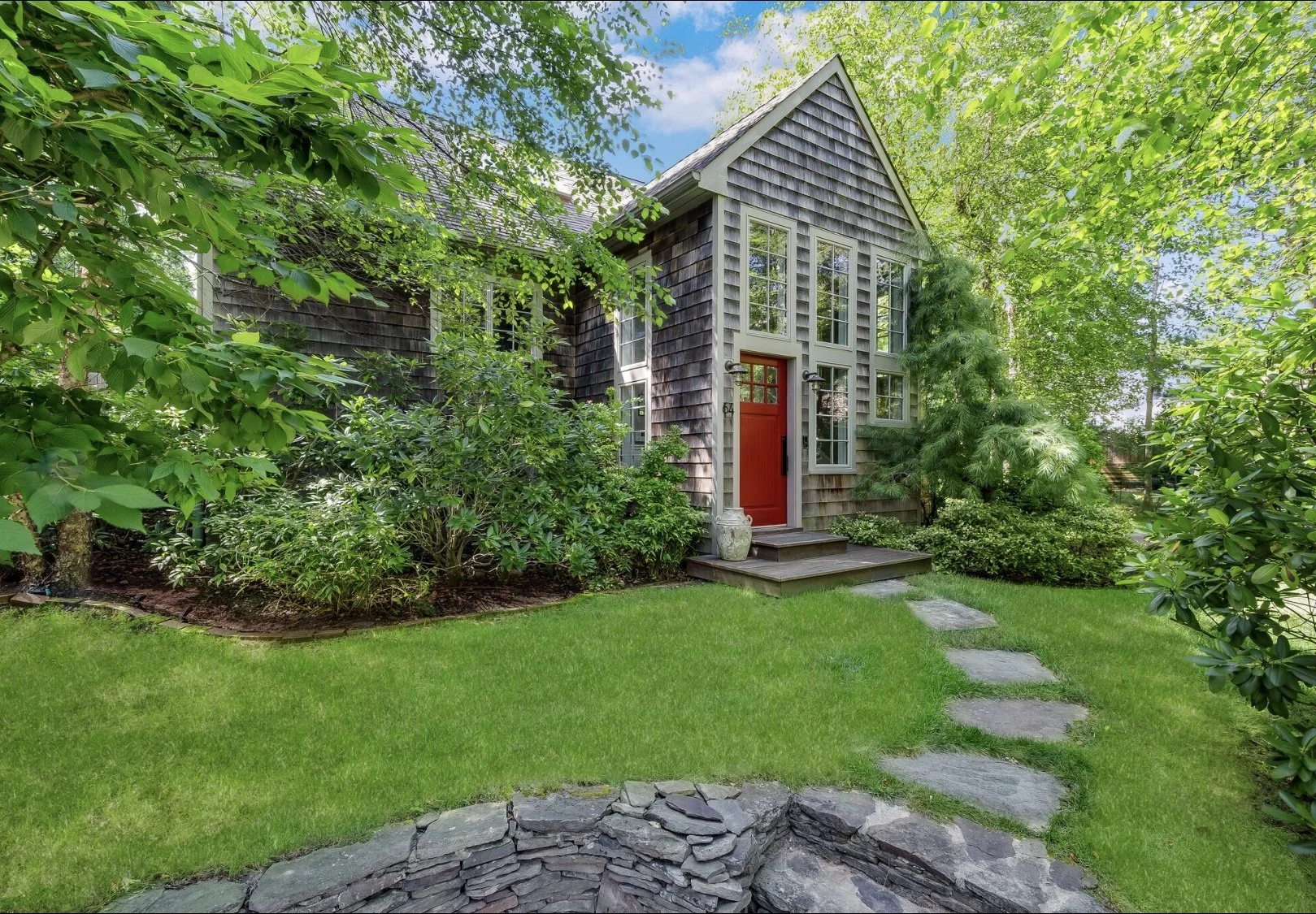 Cedar-clad cottage entrance with red front door, tall windows, and stone path set within lush, mature landscaping