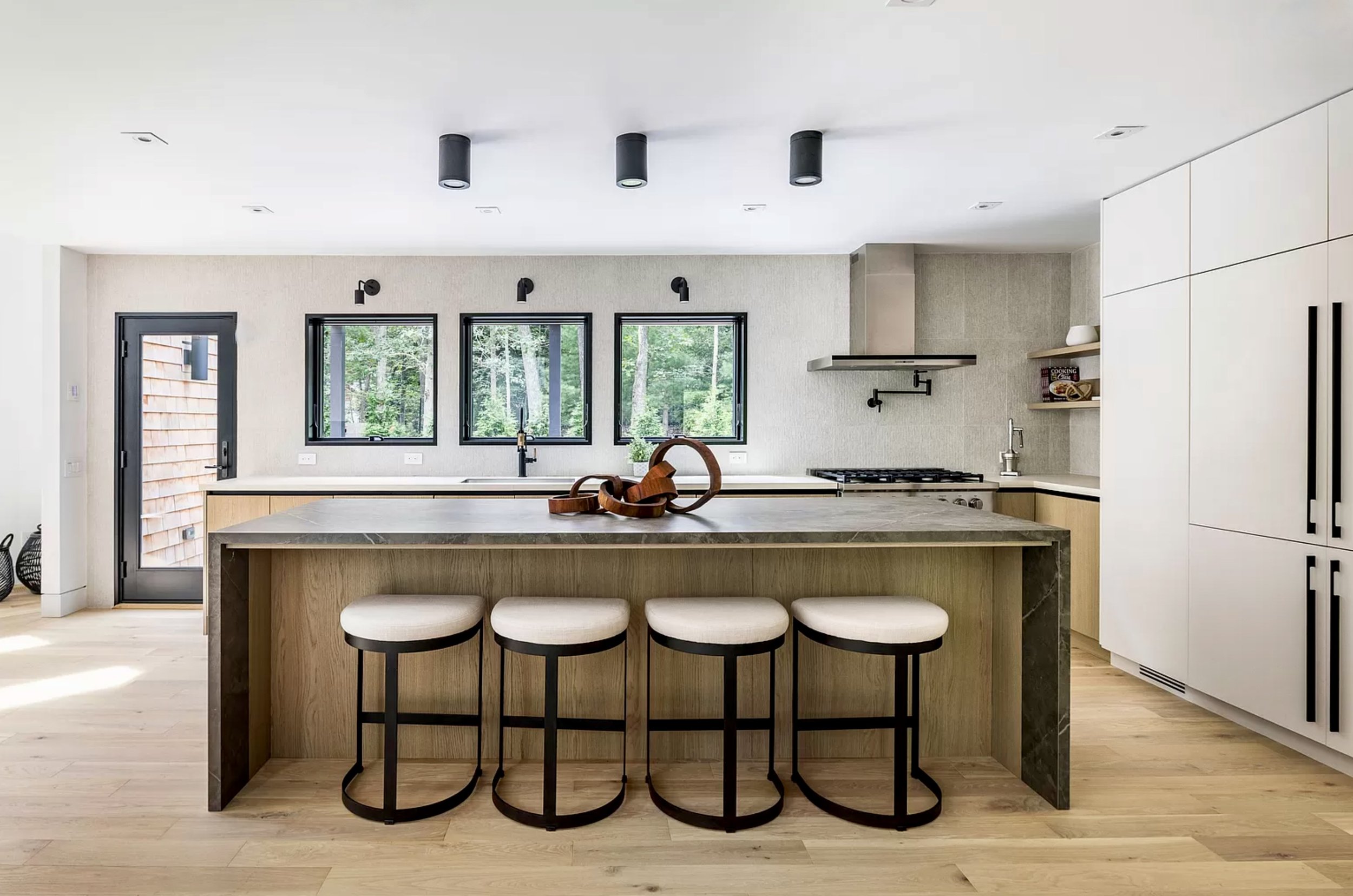 Bright modern kitchen featuring a stone island with bar stools, minimalist white cabinetry, black fixtures, and a wall of windows overlooking trees