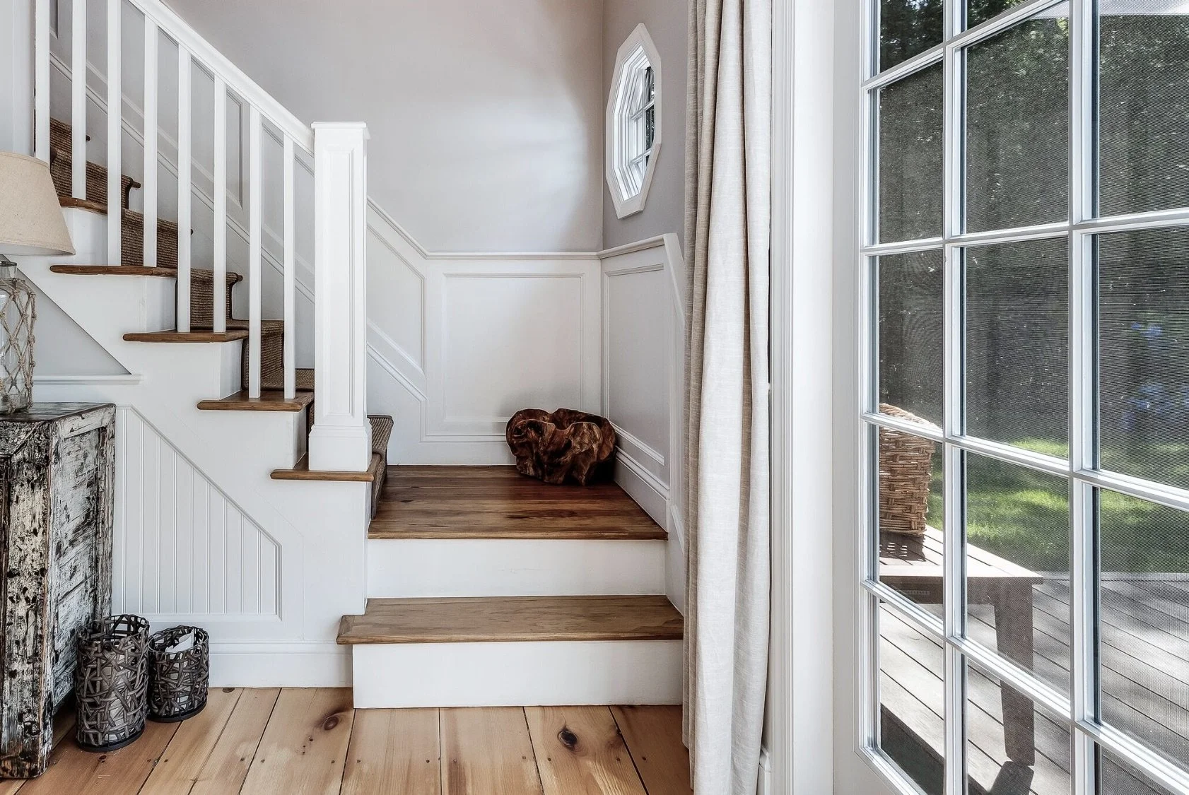 Sunlit stair hall with white wainscoting, wood treads, and a glass door opening to an outdoor deck, combining classic detailing with natural light