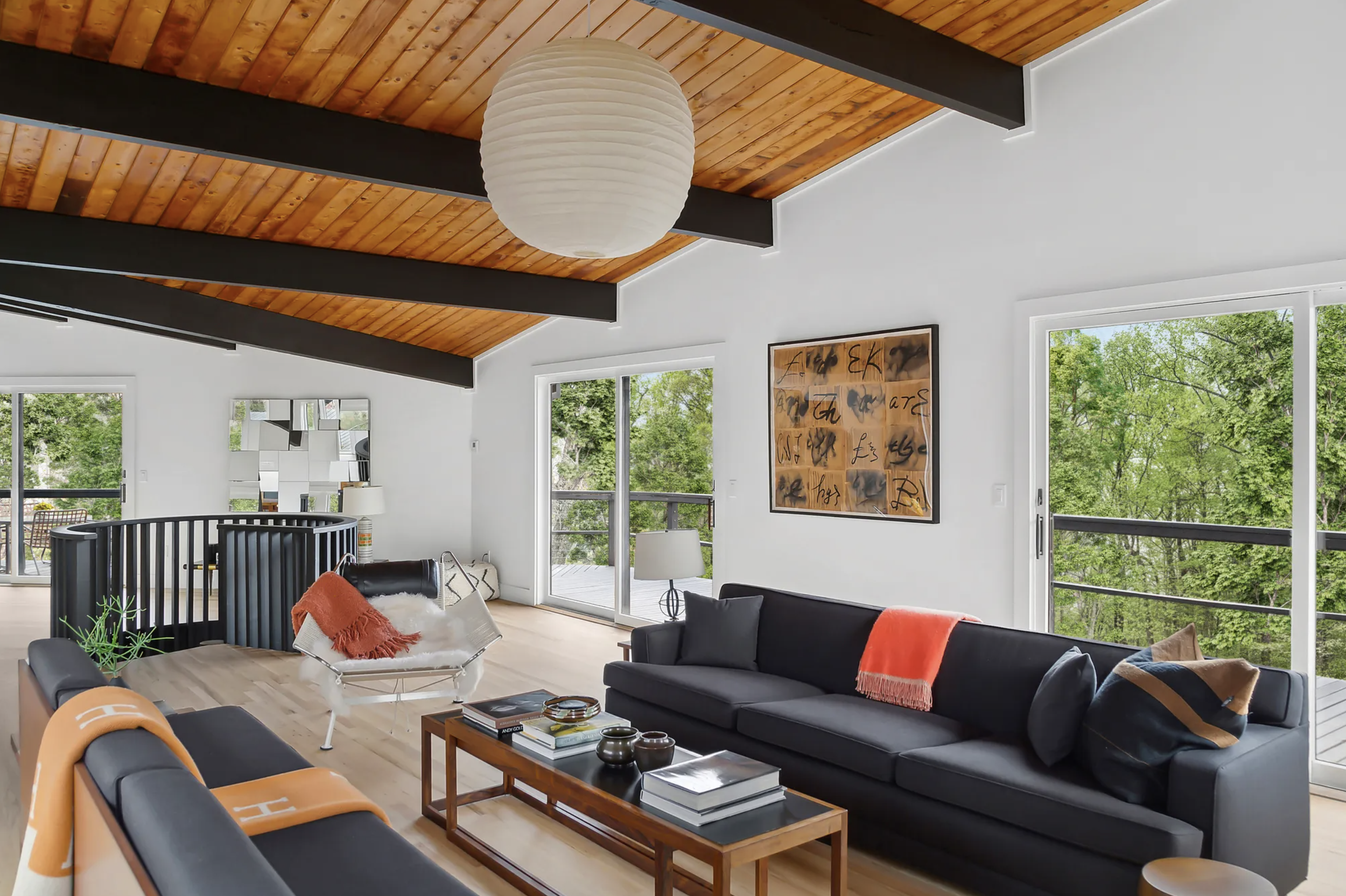Living room with vaulted ceiling, exposed beams, black sofa with warm accents, layered seating, and expansive windows framing wooded views