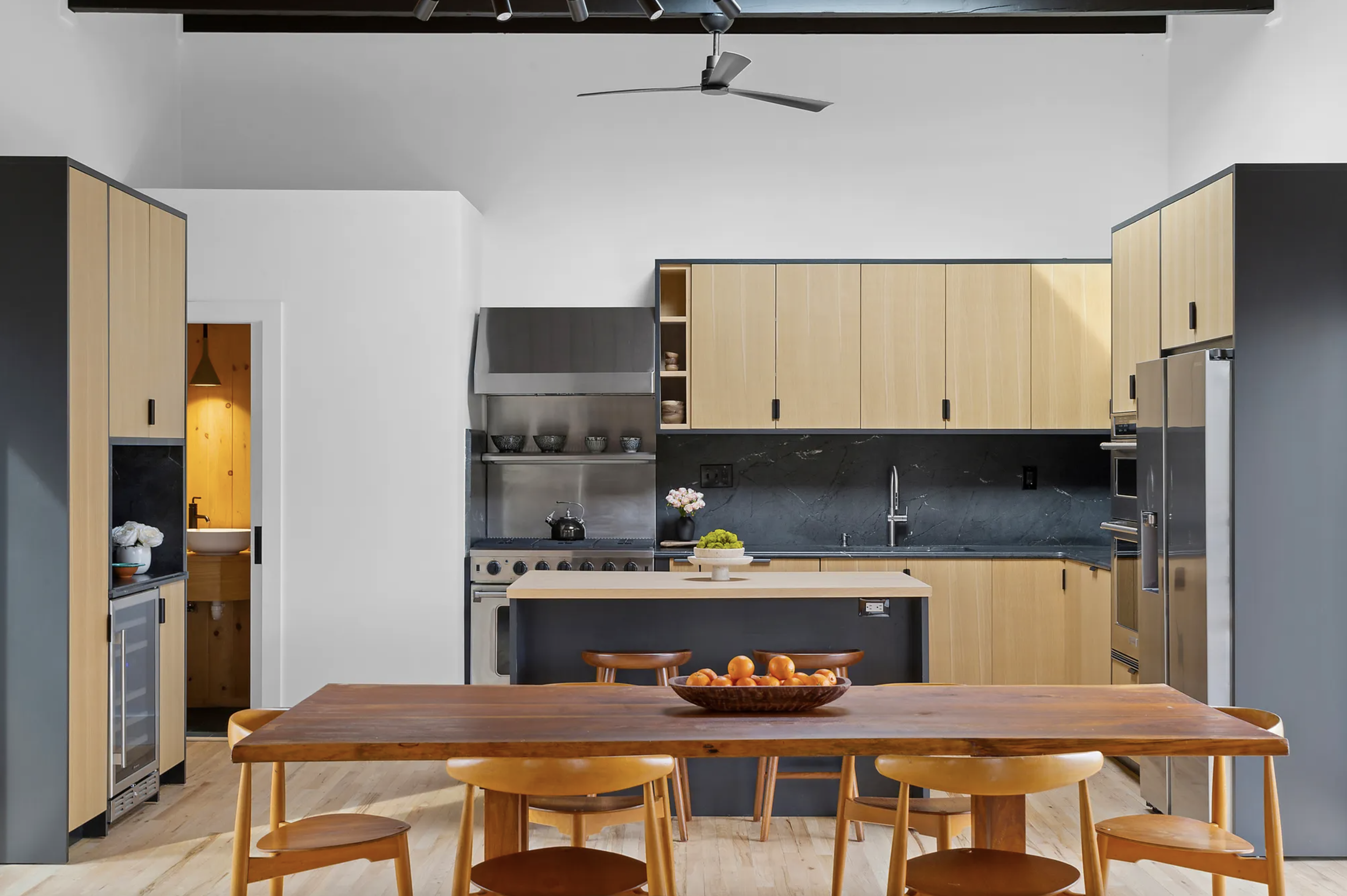 Open kitchen with light wood cabinetry, dark stone backsplash, stainless steel appliances, and a central island facing a solid wood dining table with matching chairs
