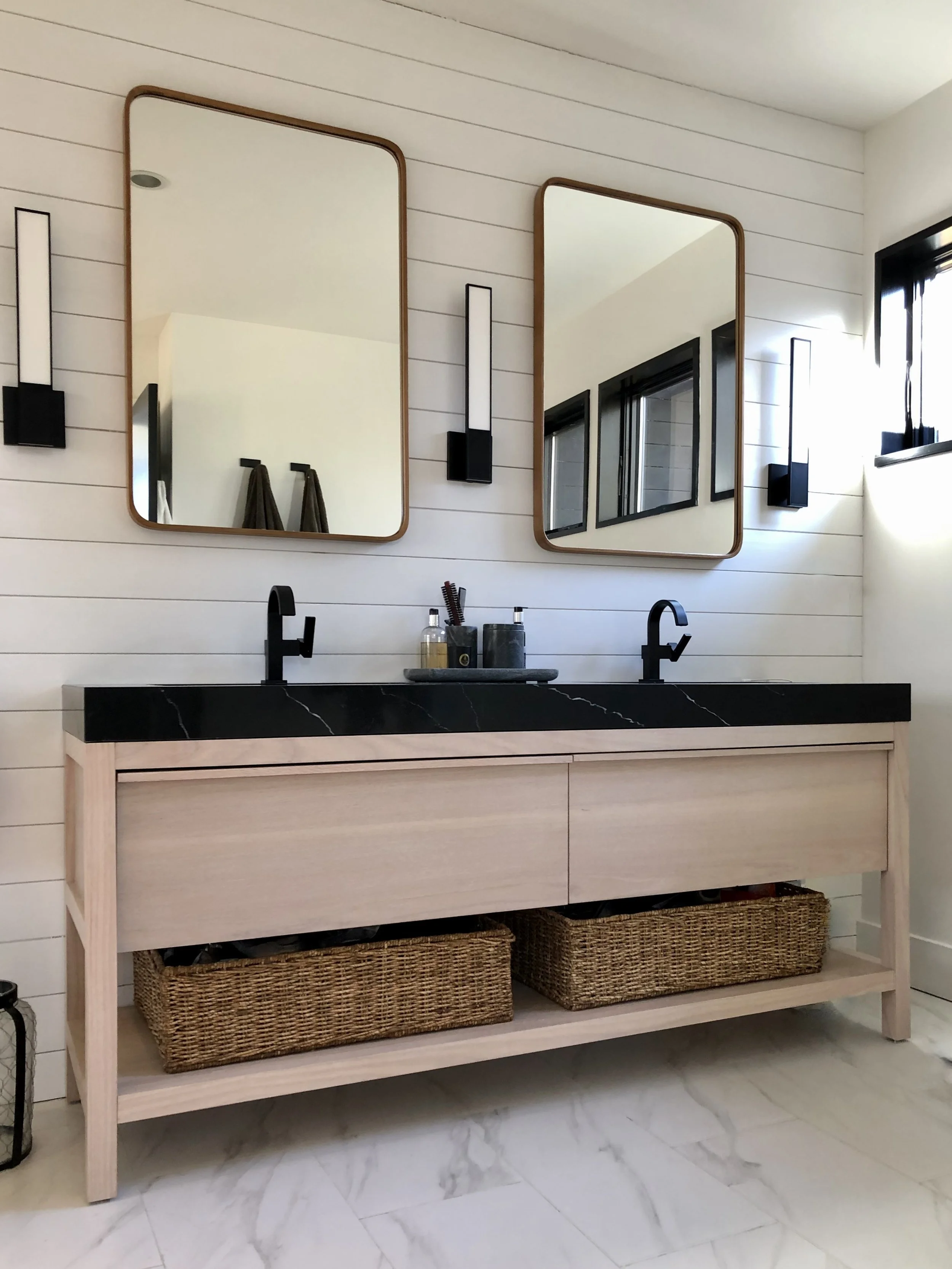 Modern bathroom vanity with light wood cabinetry, black countertop and fixtures, shiplap walls, and dual mirrors with integrated sconces