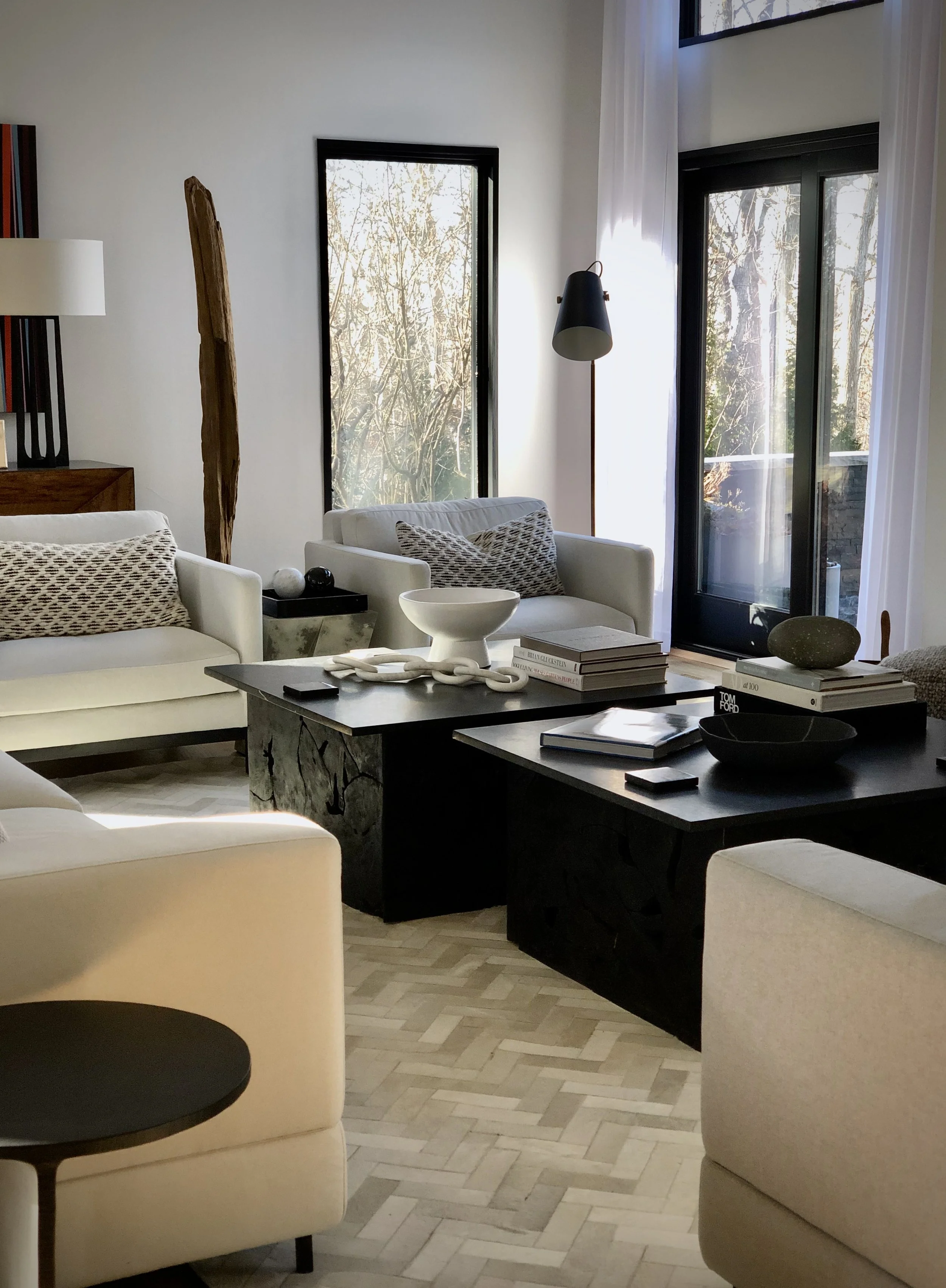 Layered living room with neutral seating, sculptural wood accents, black coffee table, and tall windows overlooking trees.