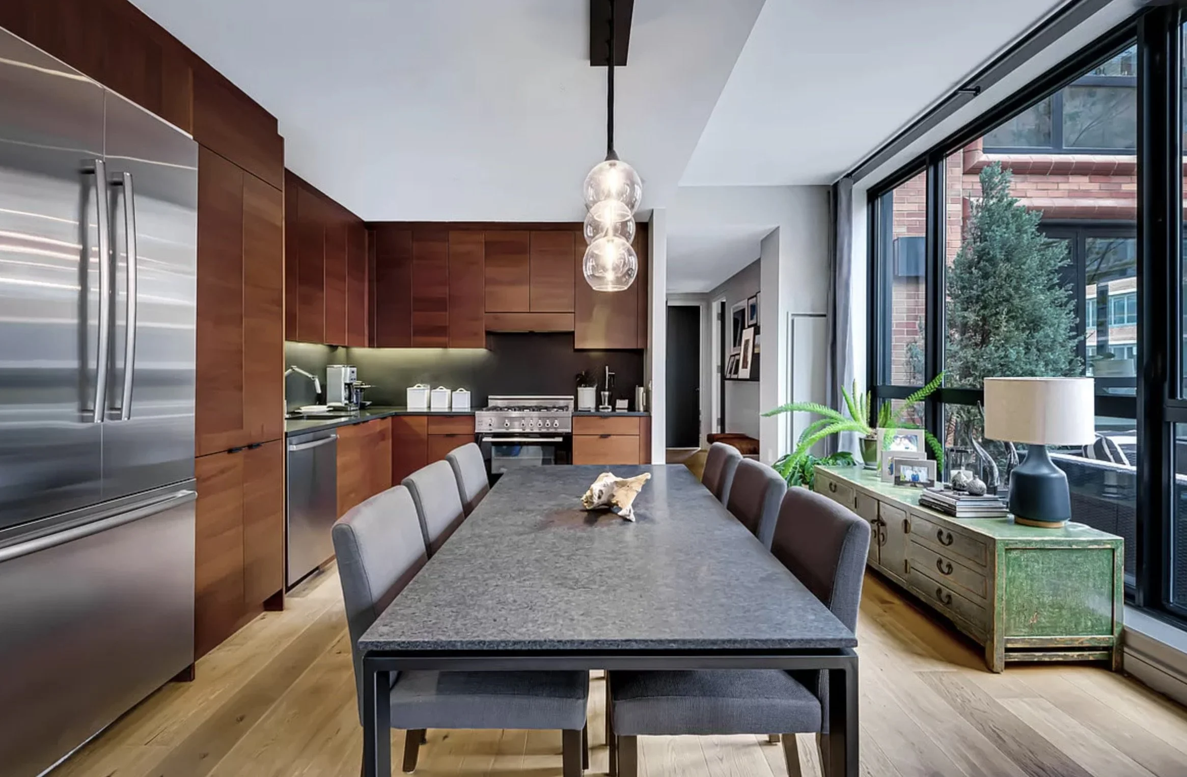 Urban kitchen and dining area with warm wood cabinetry, stone dining table, modern pendant lighting, and large windows bringing in natural light