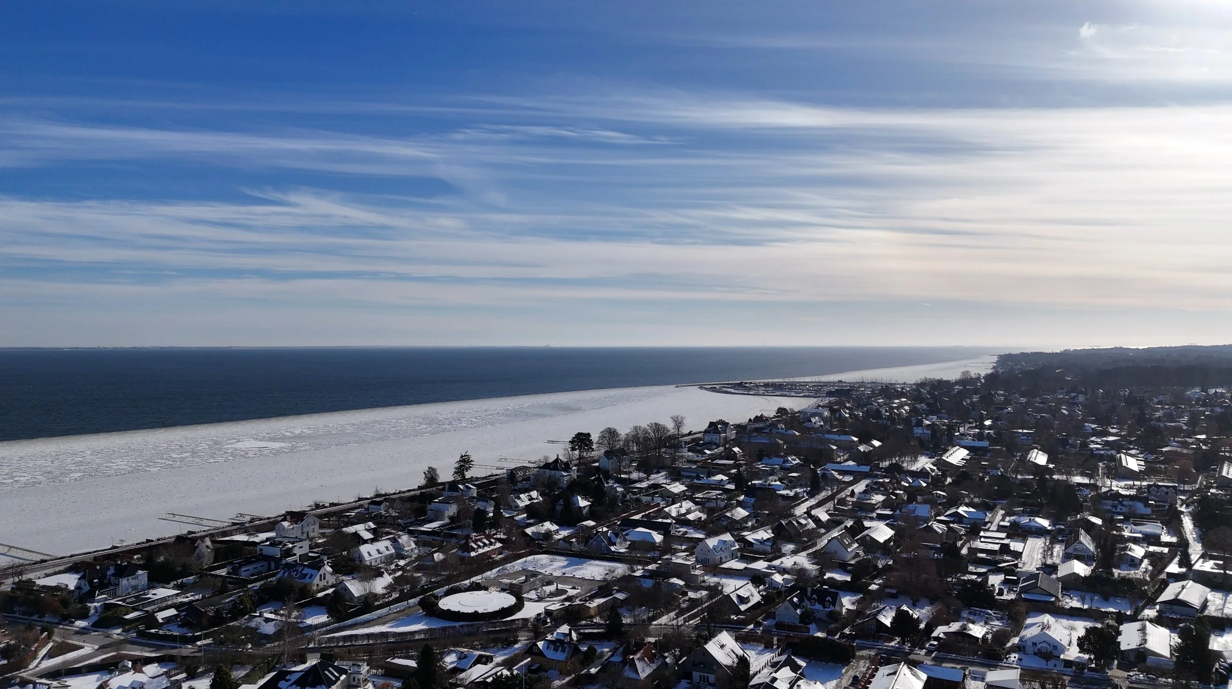 Søvindblæst vinterkyst med boliger og is langs kysten under en skyet himmel.