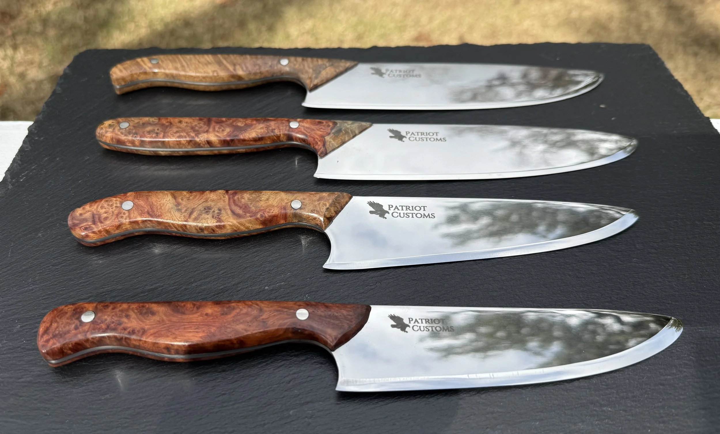 Four kitchen knives with polished blades and wooden handles, labeled 'Patriot Customs,' laid out on a black surface outdoors.