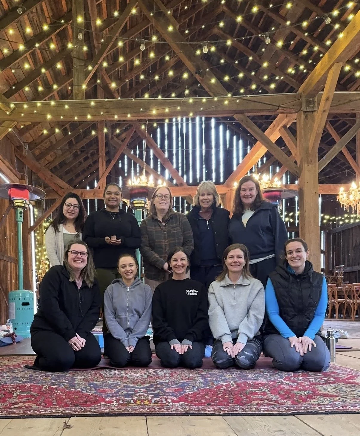 A group of ten women gathers together in a rustic wooden venue decorated with string lights and heaters, posing for a photo inside a barn or similar structure.