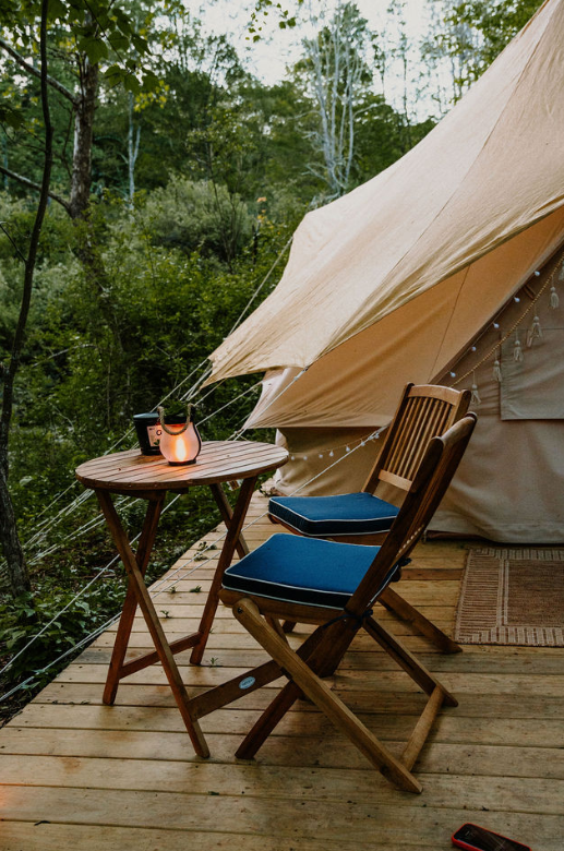 Wooden deck with two chairs and a round table outside a tent in a green wooded area during daytime, with string lights and a lantern on the table.