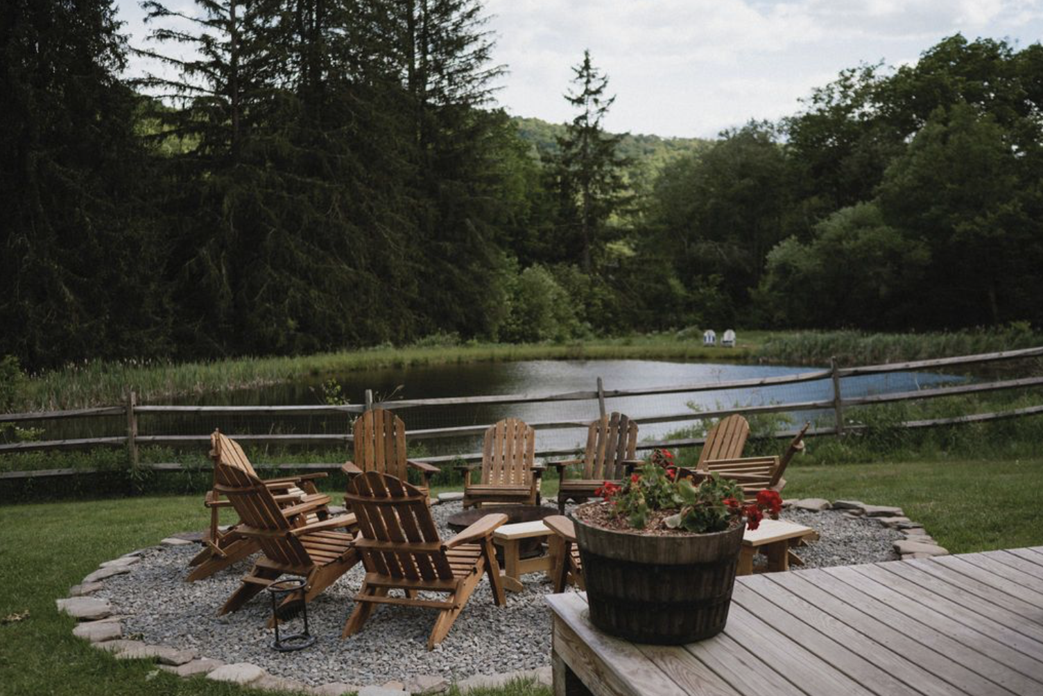 A backyard scene featuring a fire pit surrounded by wooden Adirondack chairs and a potted plant with red flowers. A wooden fence runs along a body of water, with dense trees in the background.