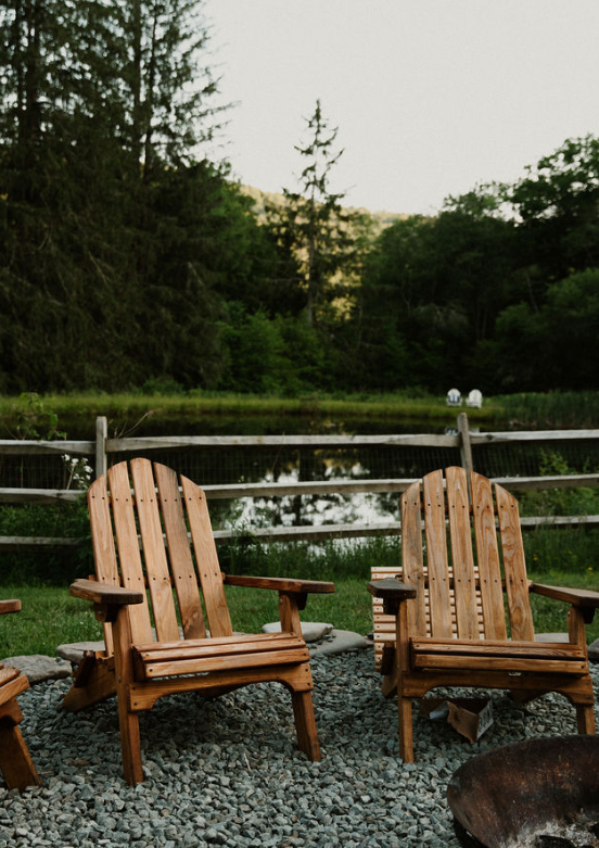 Two wooden Adirondack chairs facing a pond and surrounded by lush greenery with trees and a fence in the background.
