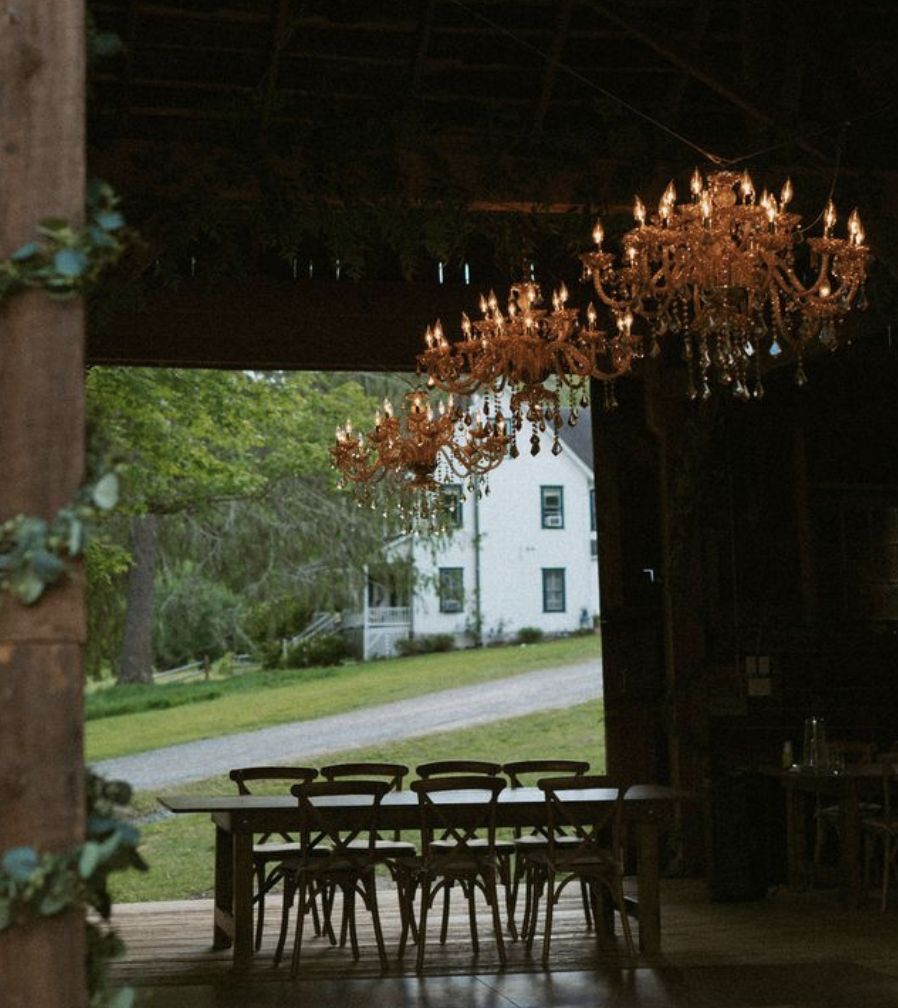 Interior of a rustic barn with chandeliers, a long wooden table, and chairs, looking outside to a green yard and white house.