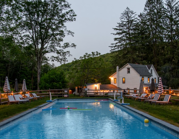 Outdoor swimming pool with floating toys and yellow ball, surrounded by sun loungers with striped cushions and umbrellas, house and trees in the background at dusk.