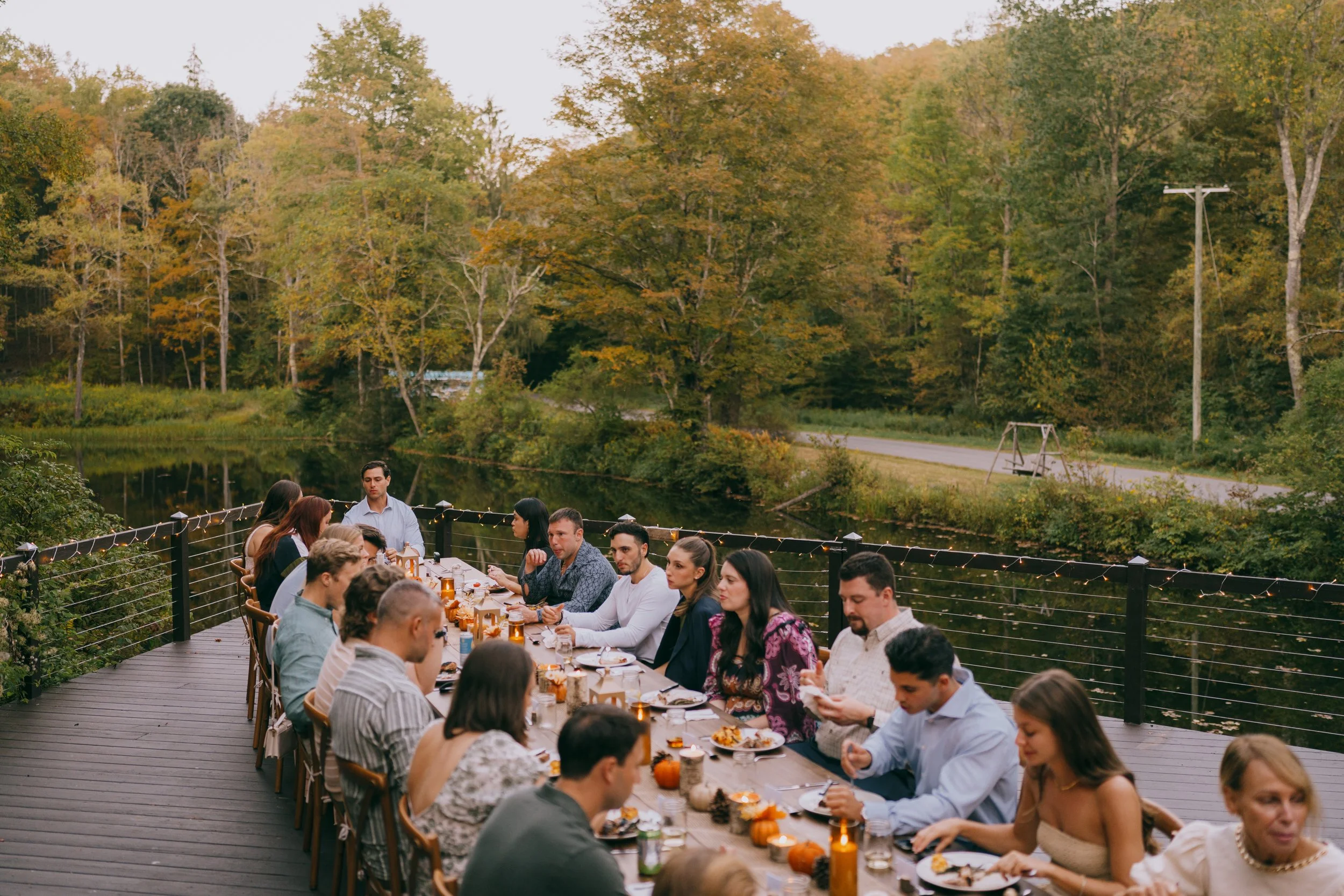 People gathered around a long outdoor dinner table by a lake, surrounded by autumn trees, with string lights and small pumpkins decorating the table.