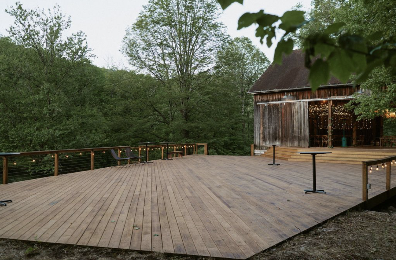 A spacious outdoor wooden deck with string lights, surrounded by lush green trees, attached to a rustic wooden barn with an open side decorated with string lights.