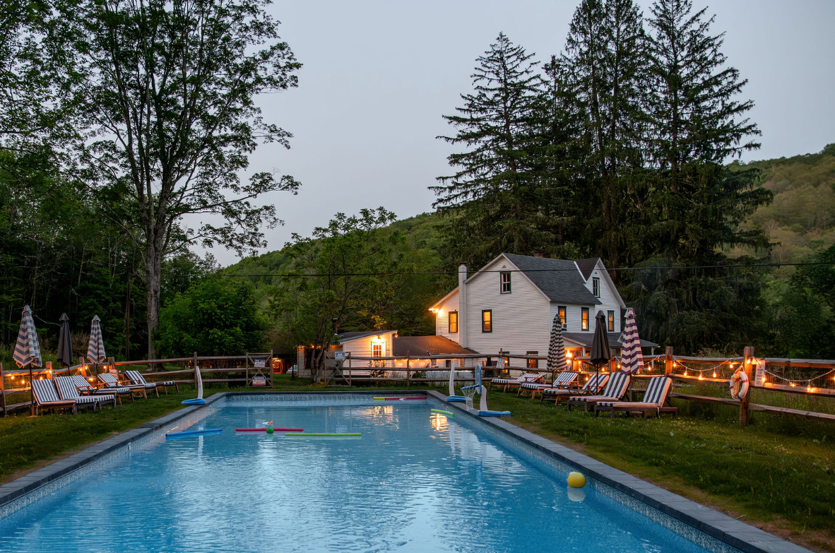 An outdoor swimming pool surrounded by lounge chairs with striped cushions and umbrellas, string lights, a white house, and lush green trees on a cloudy evening.
