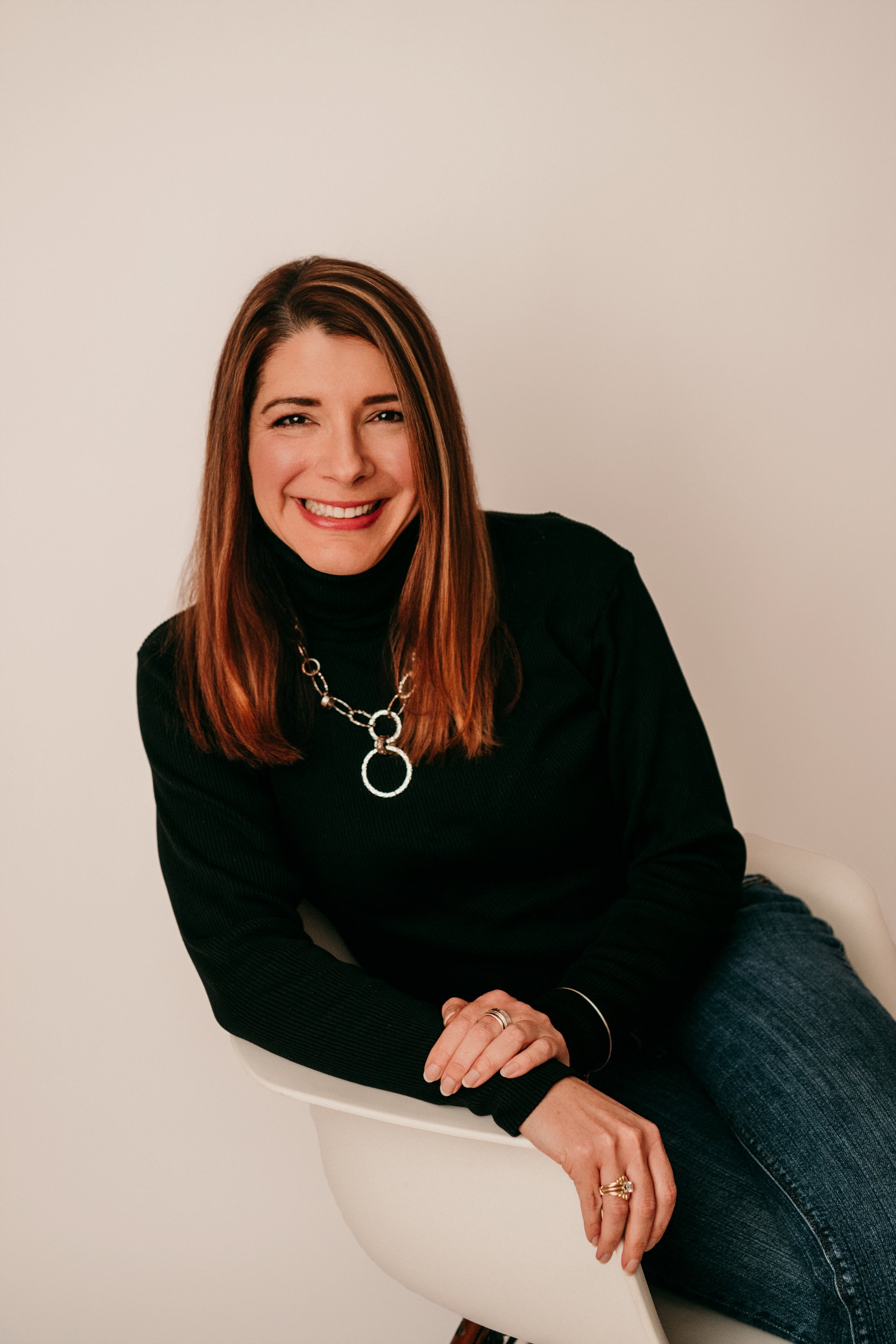 A woman with shoulder-length red hair, wearing a black turtleneck and a silver necklace, smiling while sitting in a white chair against a plain light-colored background.