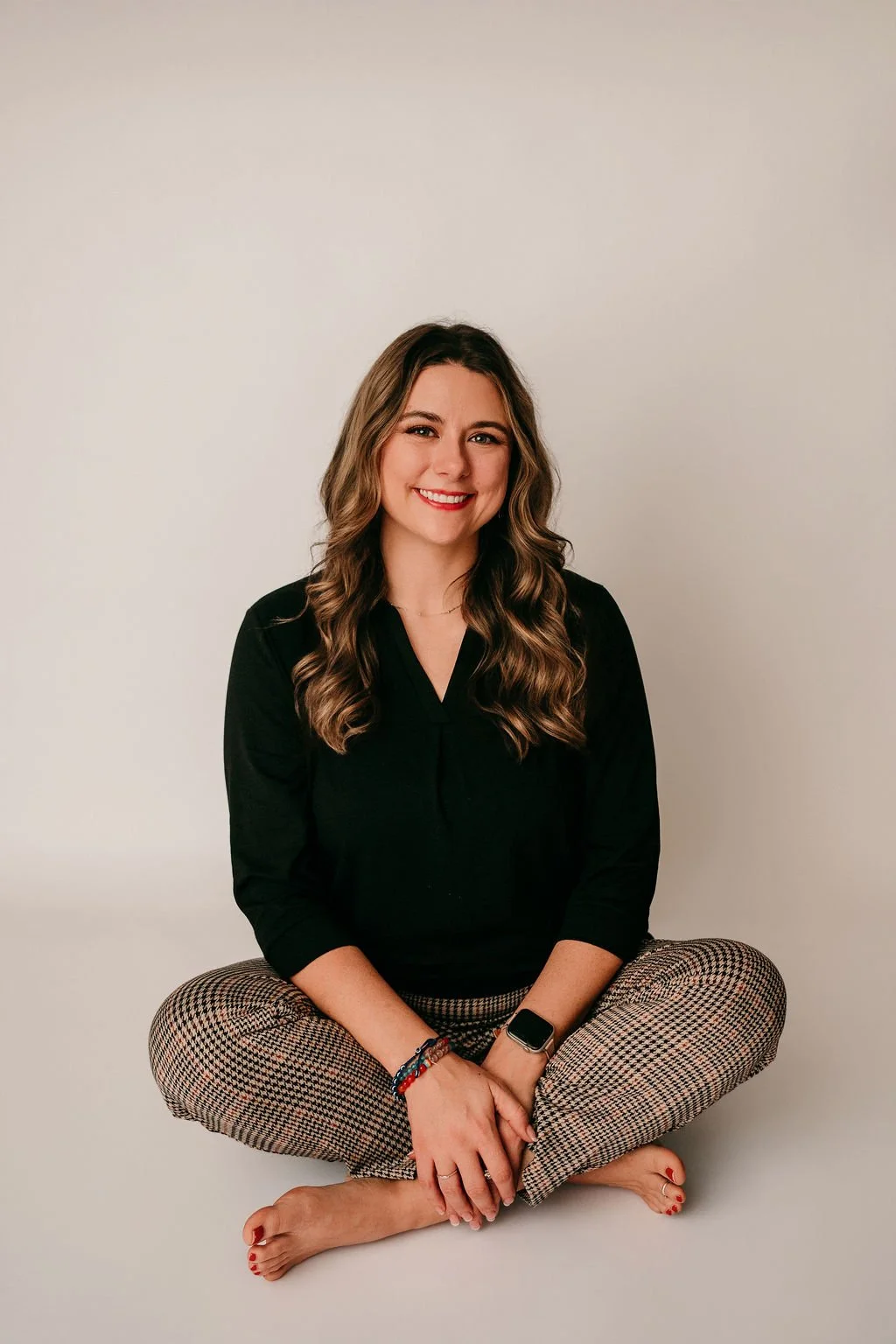 Woman with brown wavy hair, smiling, sitting barefoot cross-legged against a plain light-colored background, wearing a black top and checkered pants.