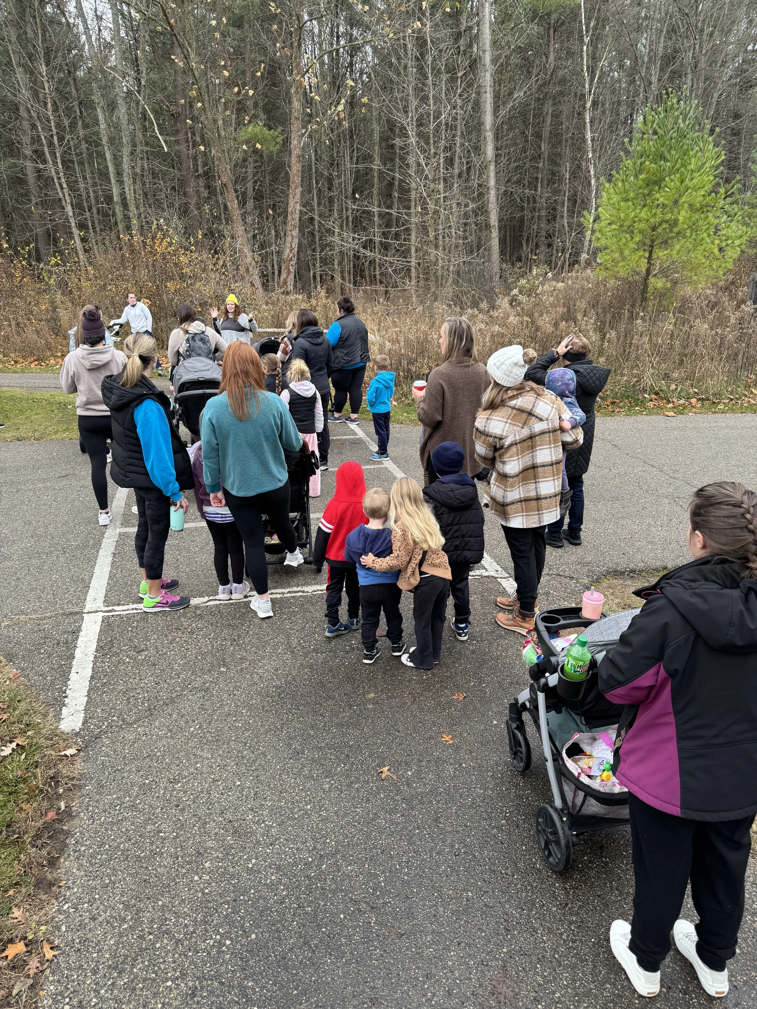 A group of people, including children and adults, gathered outdoors near a wooded area, possibly for an event or activity.