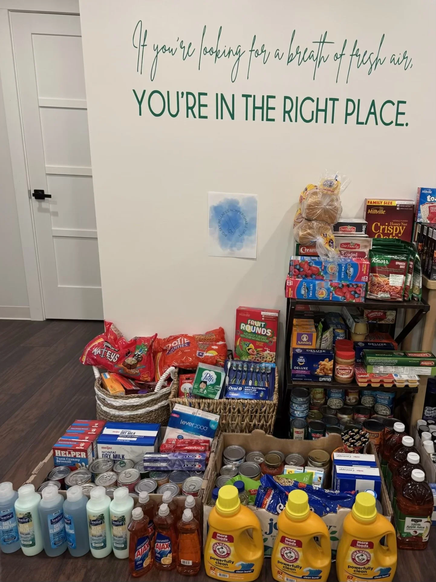 A display of groceries including canned goods, laundry detergent, snacks, milk, and personal care items, with a wall in the background that has a motivational quote about fresh air and being in the right place.