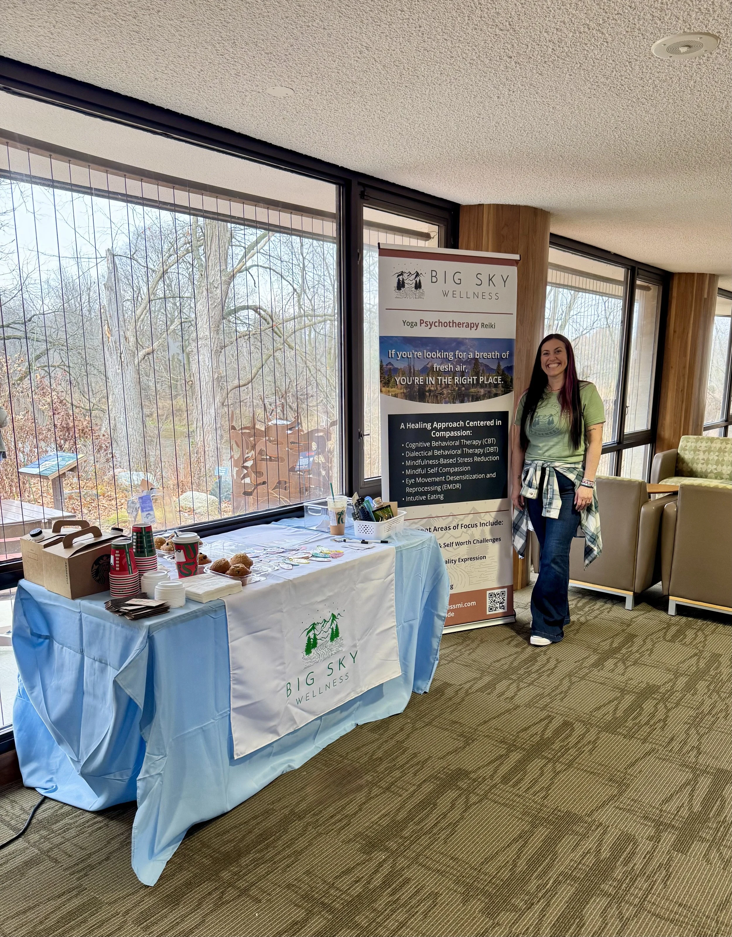 A woman standing next to a promotional banner for Big Sky Wellness, near a table with coffee cups, snacks, and informational materials, inside a room with large windows showing leafless trees outside.