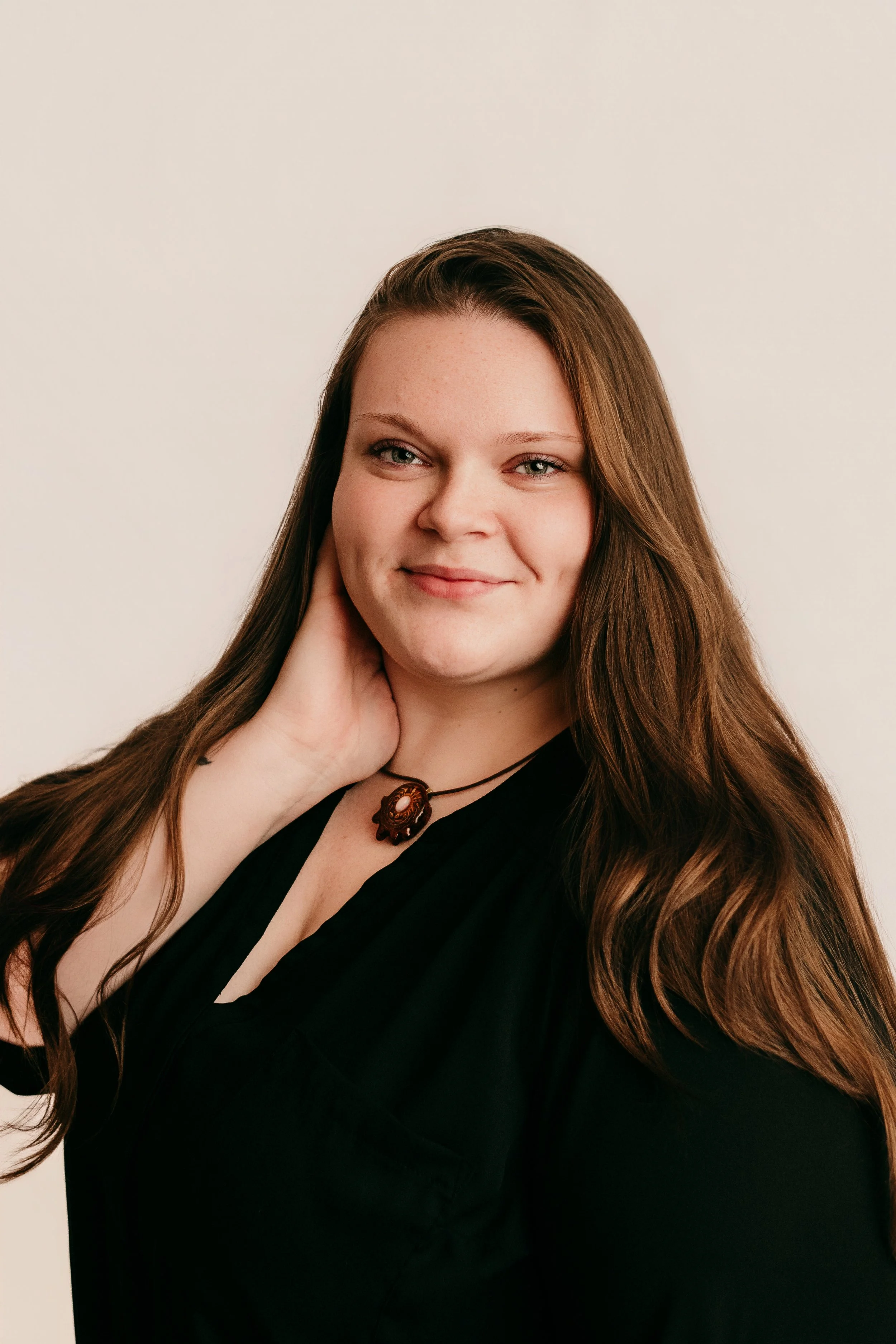 Portrait of a woman with long wavy brown hair, wearing a black top, a necklace with a large pendant, smiling slightly, with her hand resting on her neck against a plain light-colored background.