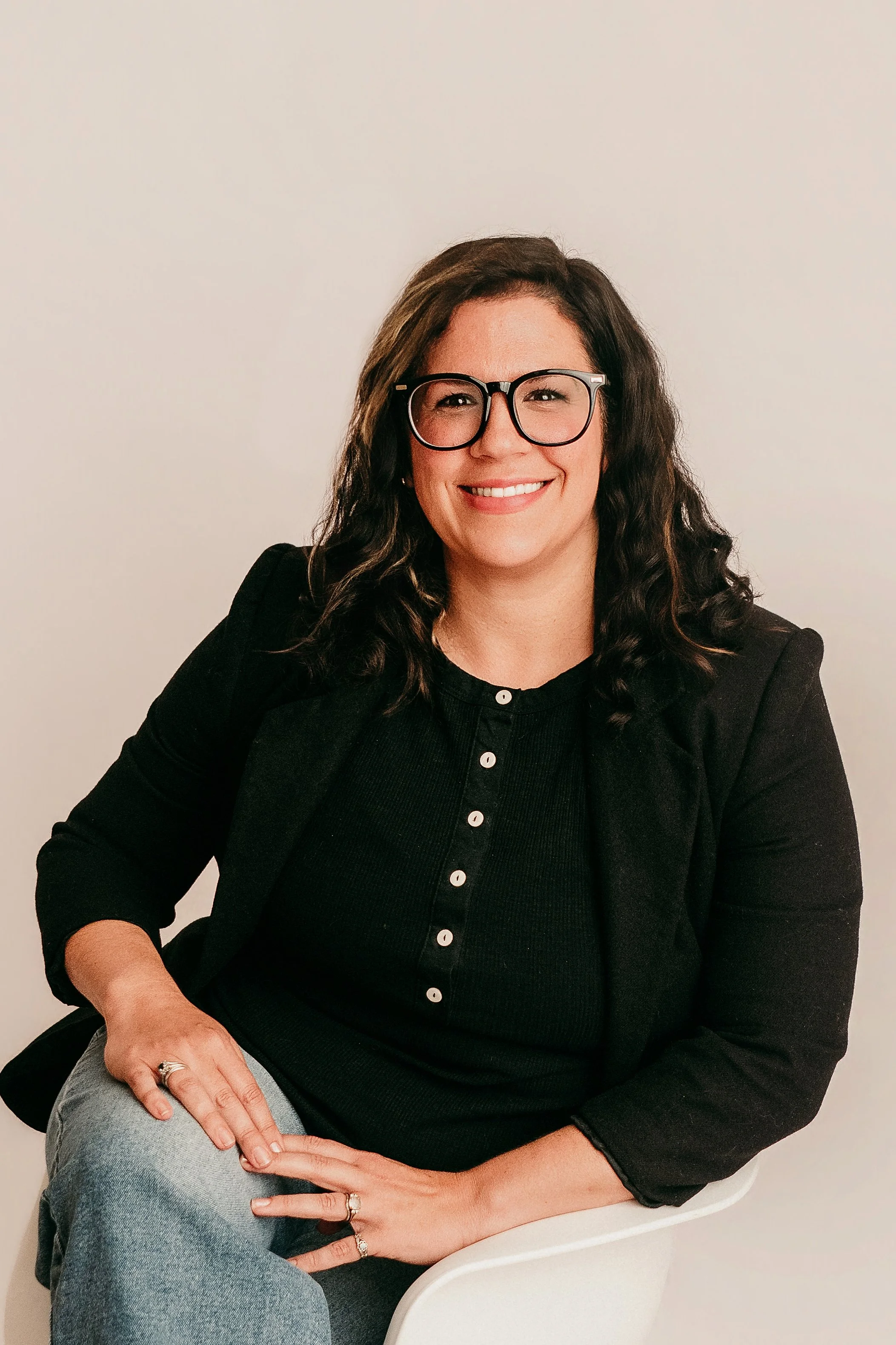 A woman with glasses and long dark hair smiling, sitting on a white chair against a plain off-white background. She is wearing a black button-up top and gray jeans.