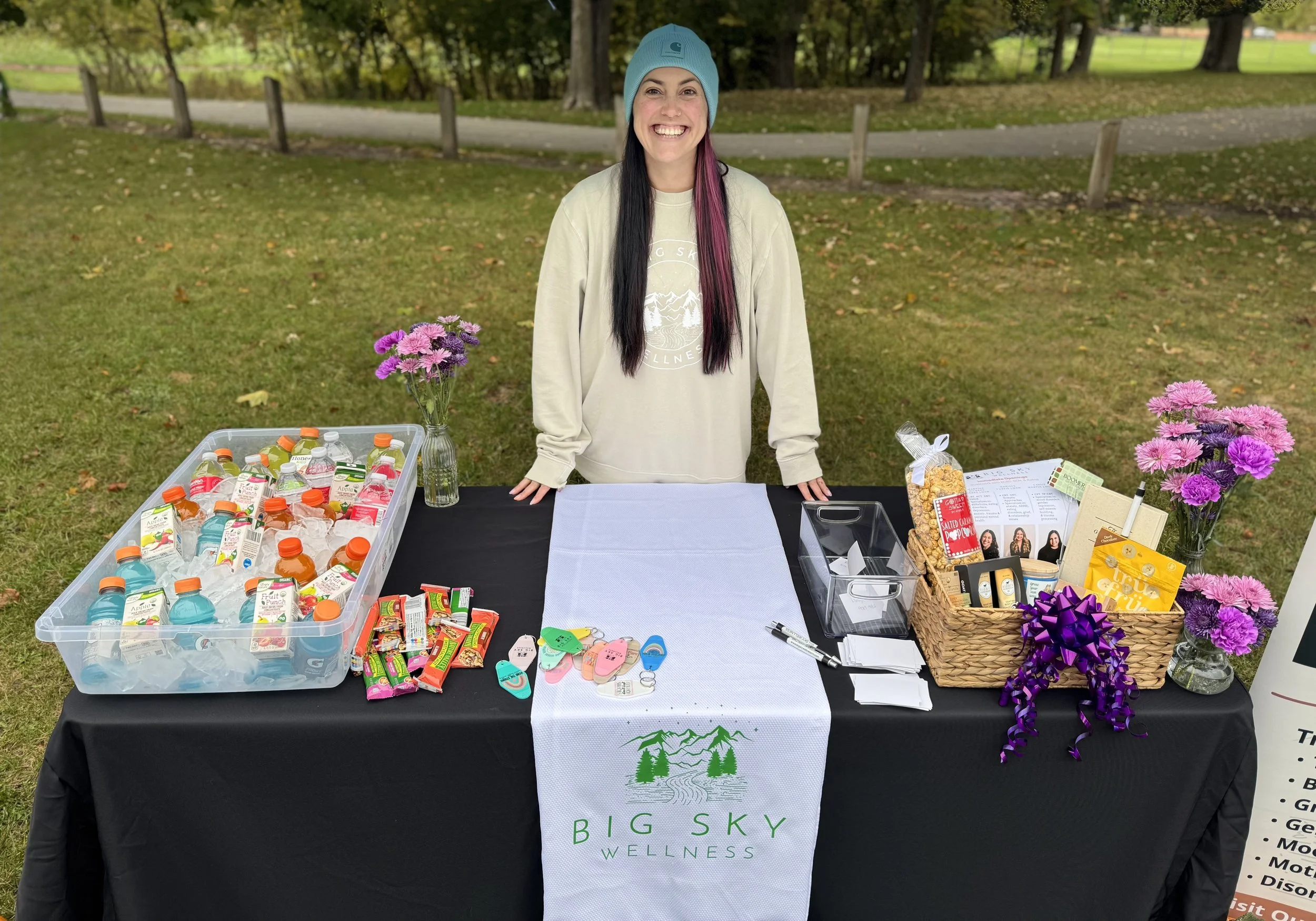 A young woman with long dark hair and purple streaks, wearing a blue beanie and beige sweatshirt, stands behind a table with various wellness items including bottled drinks, snacks, flyers, and flowers in vases, at an outdoor event in a park.