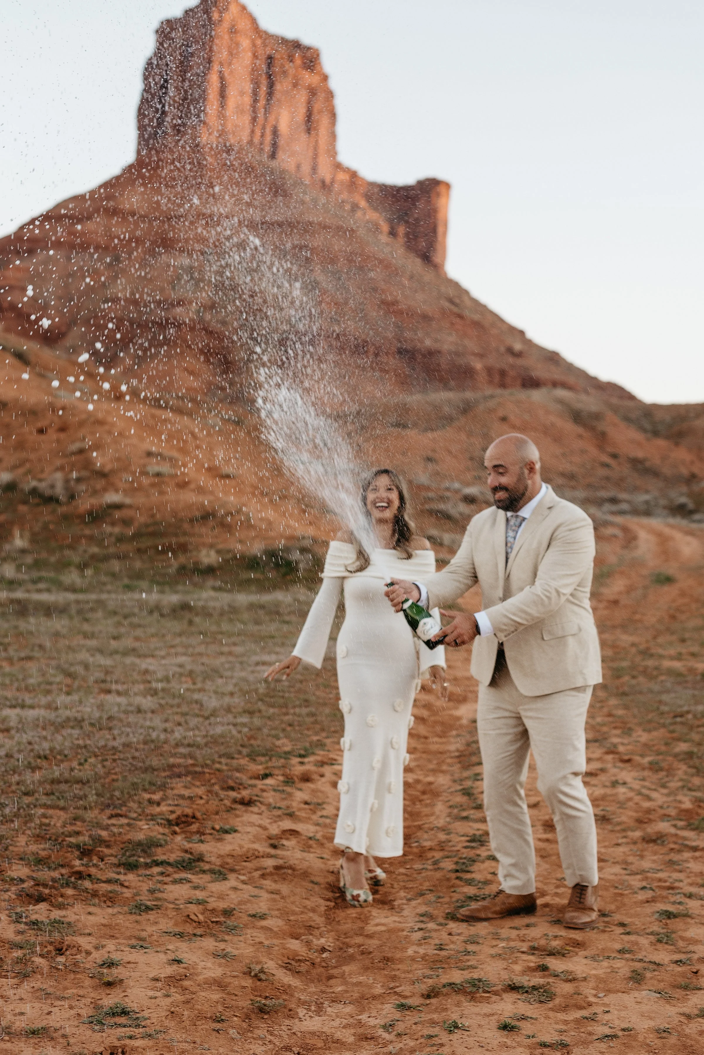 A groom is spraying a champagne bottle and a bride is next to him excitedly watching. they are standing in a red rock desert and the champagne is spraying above their heads.