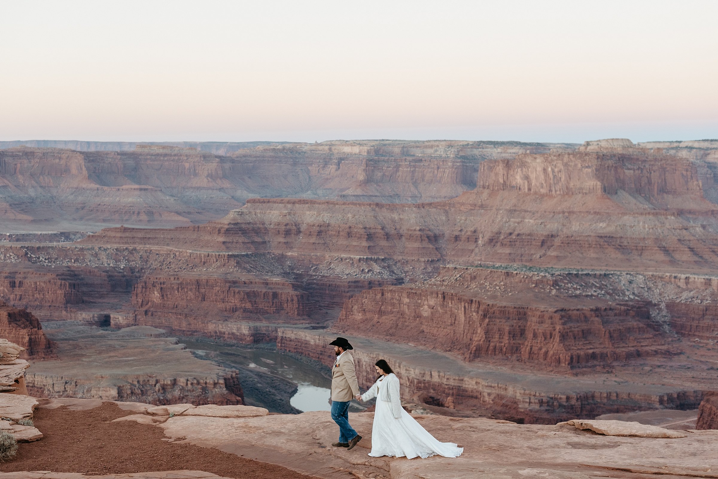 A bride and groom are walking along a cliff side. There is a large red rock canyon in the background and the colorado river. The groom is wearing a cowboy hat and holding the bride's hand.