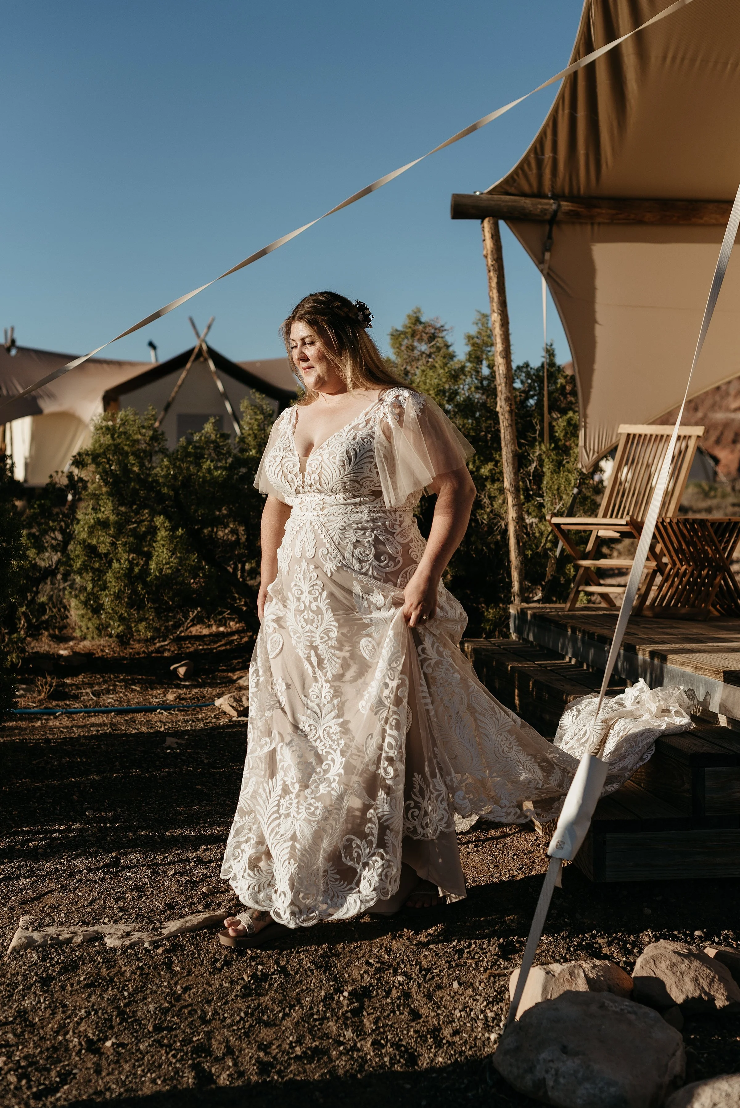 A bride is walking out of a canvas tent with more tents in the background. The sun is rising and the sky is very blue. She is holding her dress up and showing her tan hiking sandals.