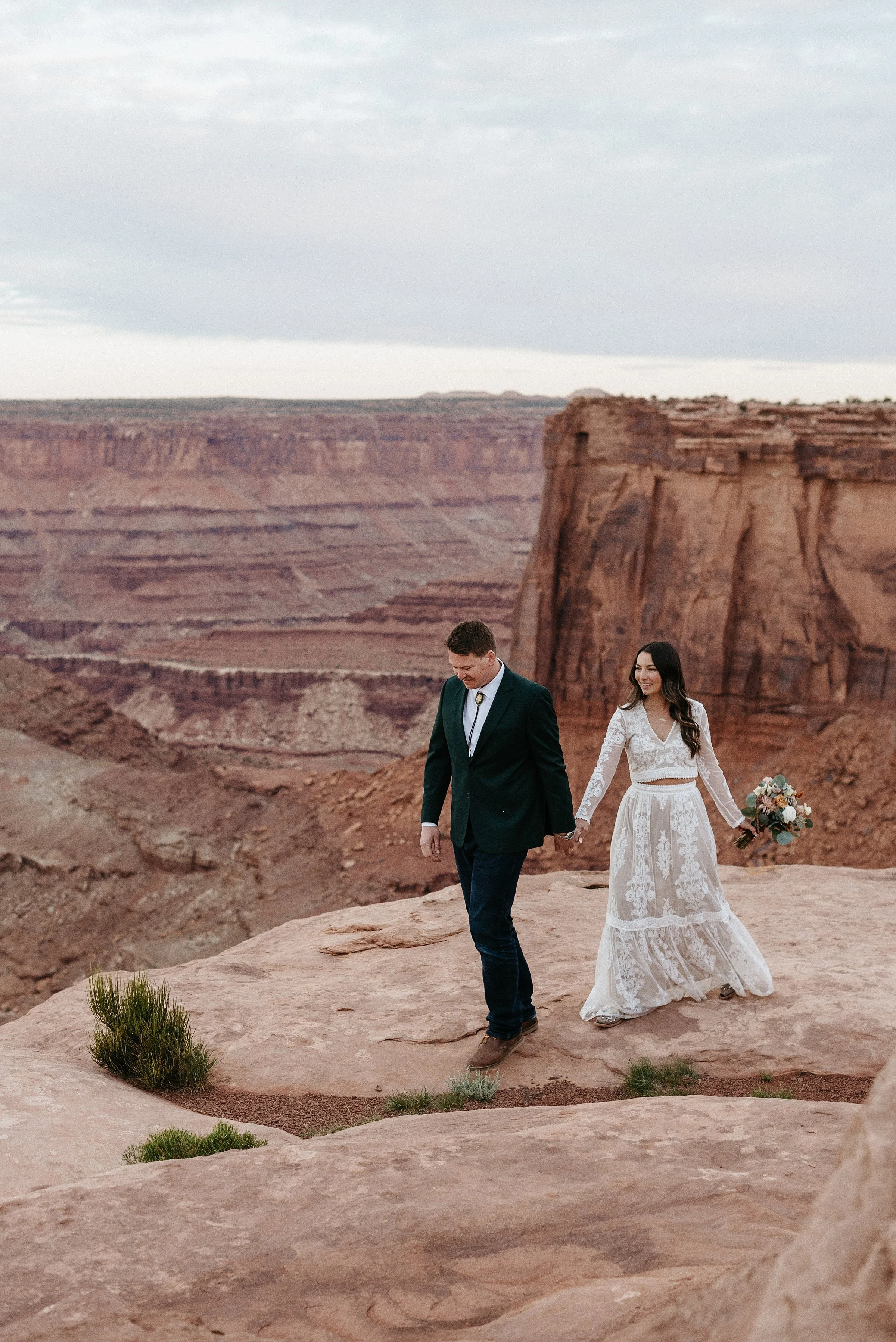 A bride and groom walk hand in hand along a red red cliff. She is wearing a two piece long sleeve lace wedding dress and holding a bouquet of flowers. He is wearing a dark green suit and bolo tie. A red rock canyon and mesa is behind them.