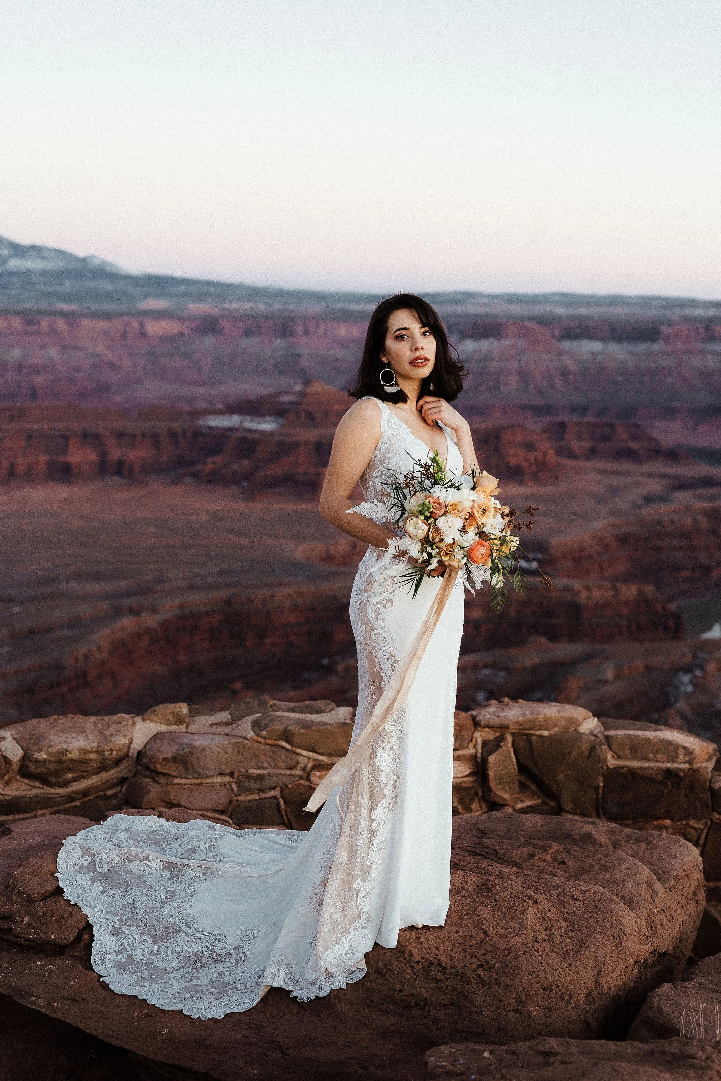 A bride is standing holding a bouquet with a large red rock canyon behind her.